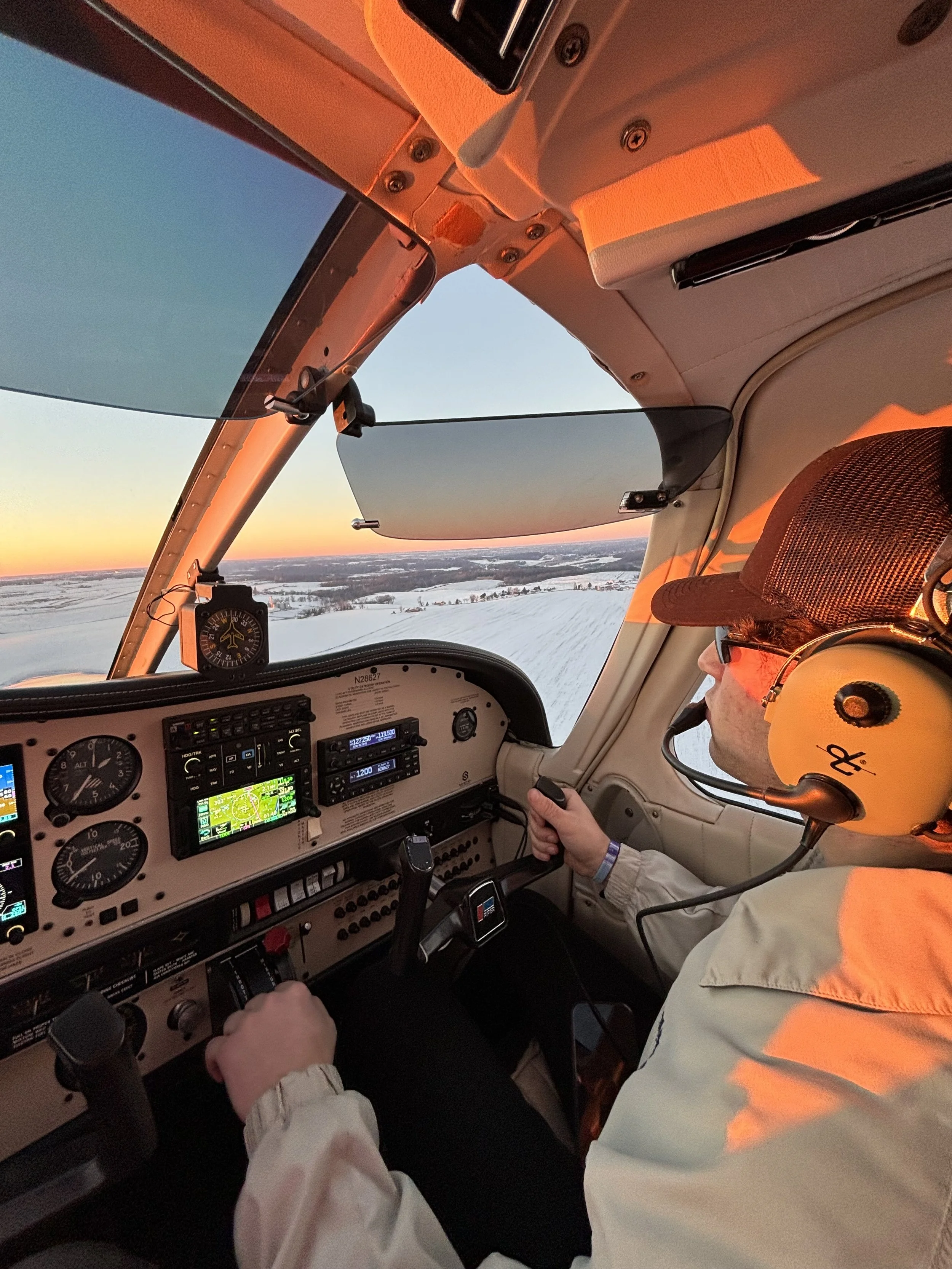 Inside a small aircraft cockpit with a pilot wearing a yellow helmet and headset, flying over a snowy landscape during sunset, with instruments and controls visible on the dashboard.