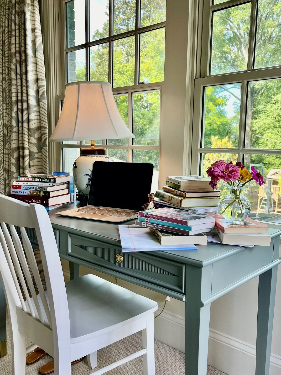 Caroline Langerman's study desk by a window with books, a laptop, a table lamp, a vase of flowers, and a white chair.