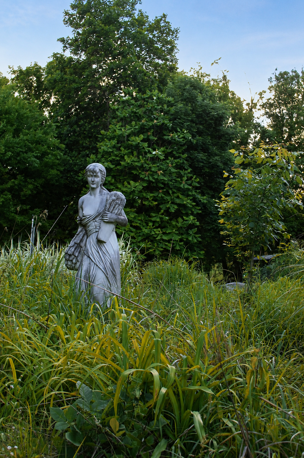 A classical statue of a woman holding a bundle of fabric, standing amidst tall grass with greenery and trees in the background under a blue sky.
