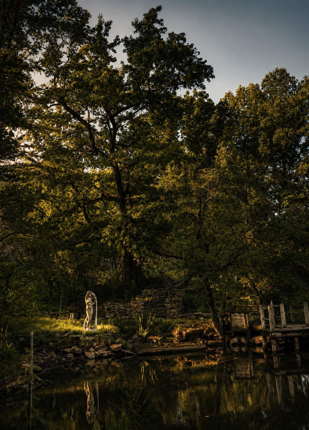 A tranquil pond surrounded by lush trees at sunset, with a white sculpture of a woman holding a staff near the water's edge and a small wooden dock on the right.