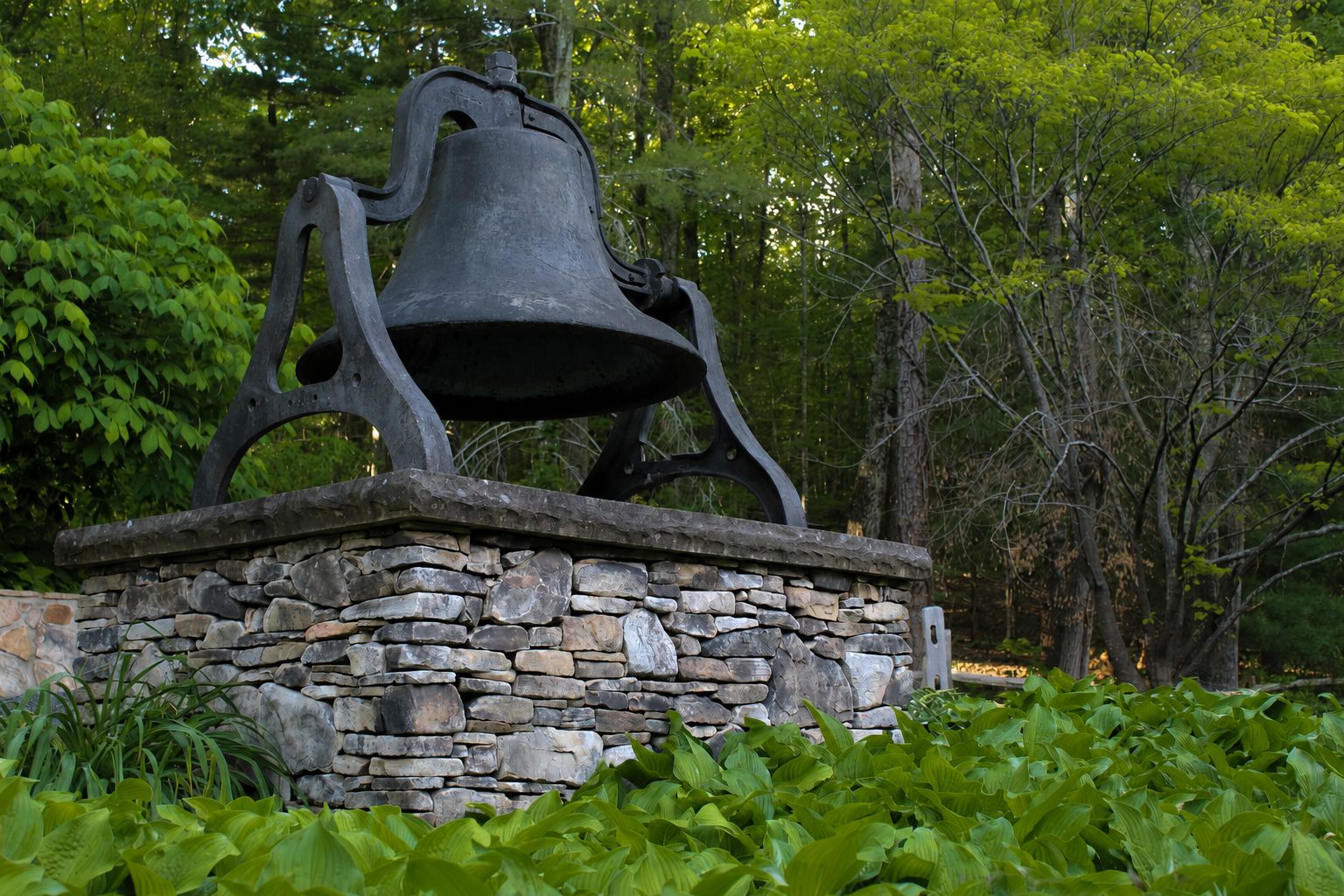 Large, antique metal bell mounted on a stone pedestal, surrounded by green plants and trees in a forest setting.