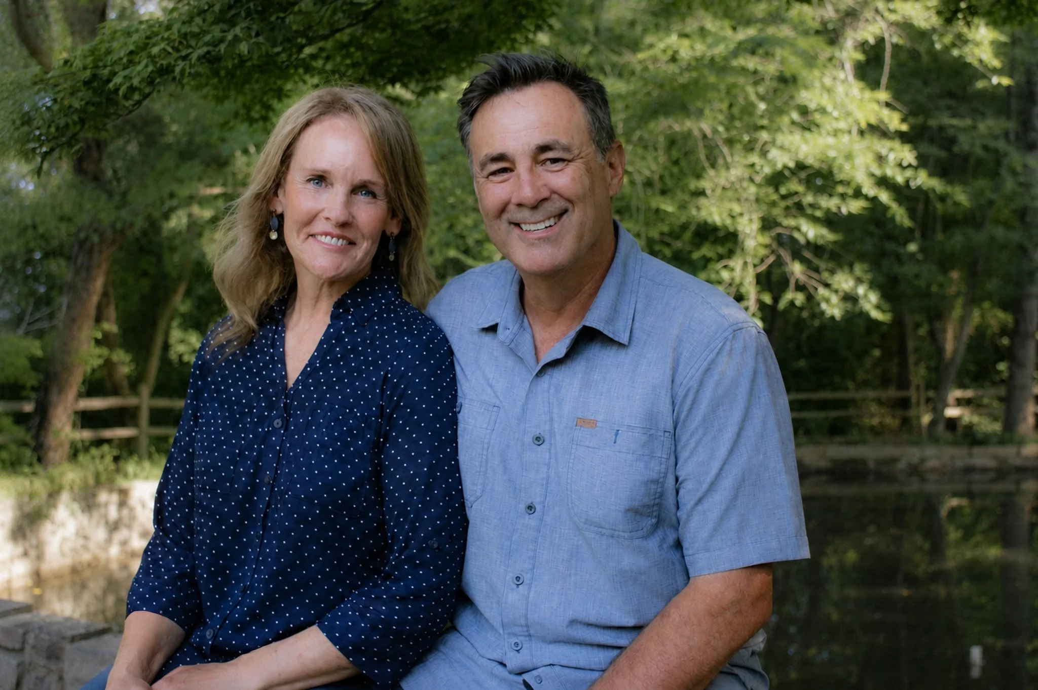 A middle-aged couple smiling outdoors, the woman with shoulder-length blonde hair wearing a blue polka-dot blouse, and the man with short dark hair wearing a light blue shirt, standing in front of green trees.