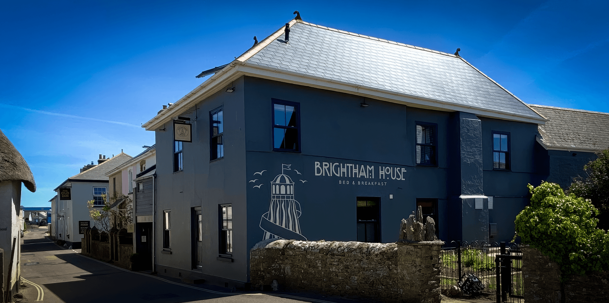 Blue building labeled 'Brightam House Bed & Breakfast' with a lighthouse illustration, situated on a street with other buildings and greenery under a clear blue sky.