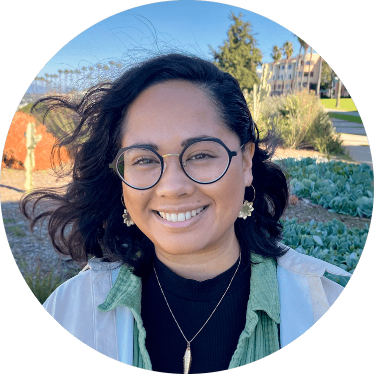 Smiling woman with glasses and earrings outdoors on a sunny day, with plants and buildings in the background.