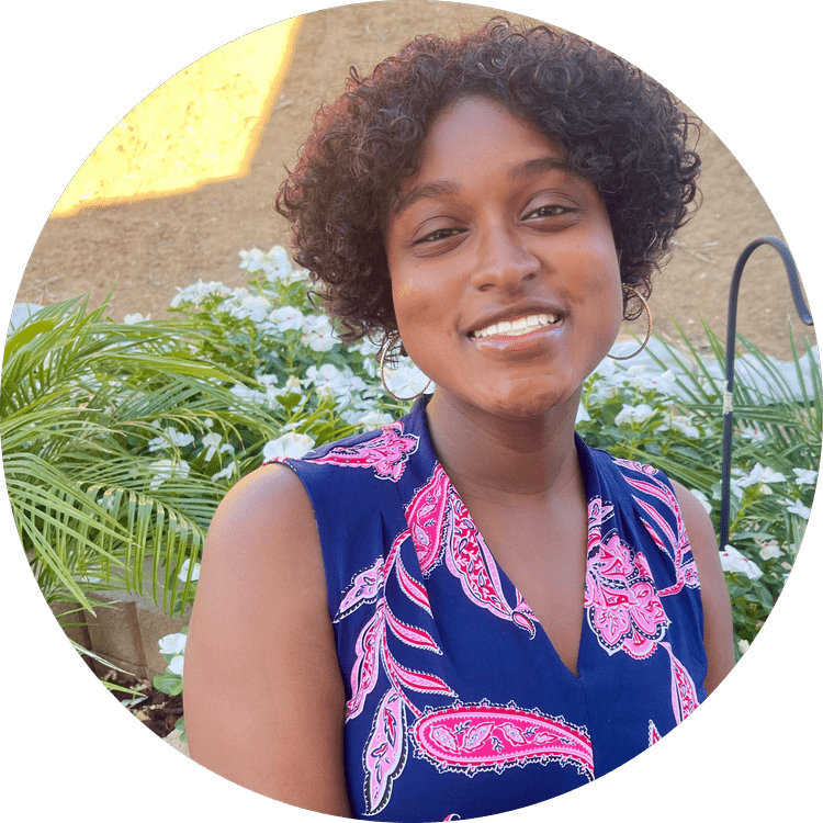 A young African American woman with short curly hair, smiling, wearing a sleeveless blue top with pink and white paisley patterns, standing outdoors in front of green plants and white flowers.