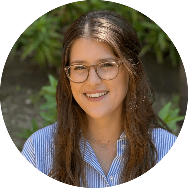 A woman with long brown hair, glasses, and a striped blue and white shirt smiling outdoors with green foliage in the background.