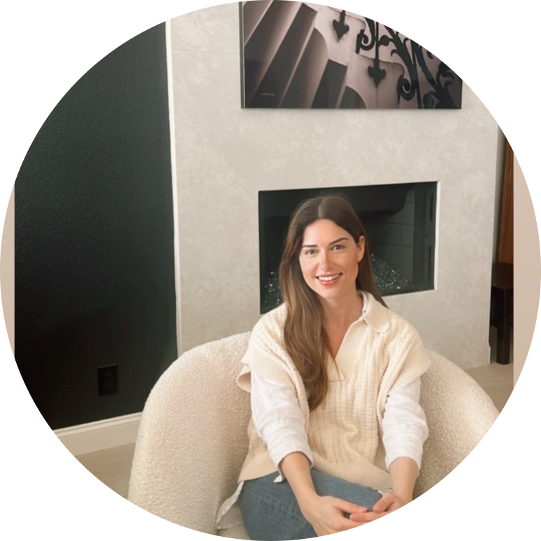 A woman sitting on a beige chair in a living room, smiling at the camera, with a fireplace and abstract artwork behind her.