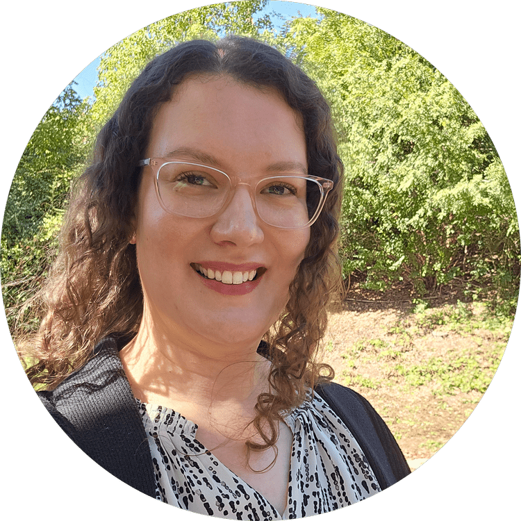 A smiling woman with curly hair, glasses, and a patterned blouse outdoors with green trees and sunlight in the background.
