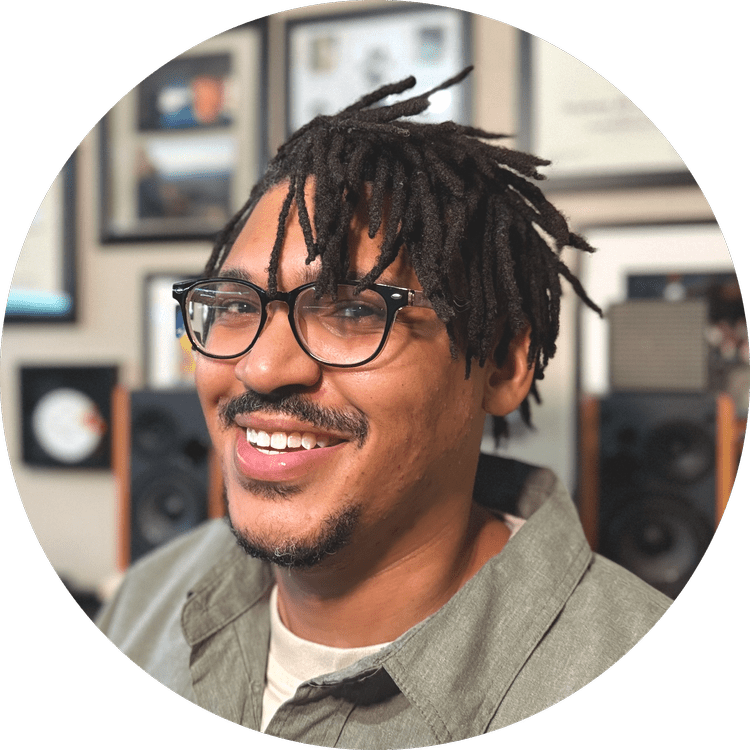 A smiling man with glasses and dreadlocks in a room with framed pictures and speakers.