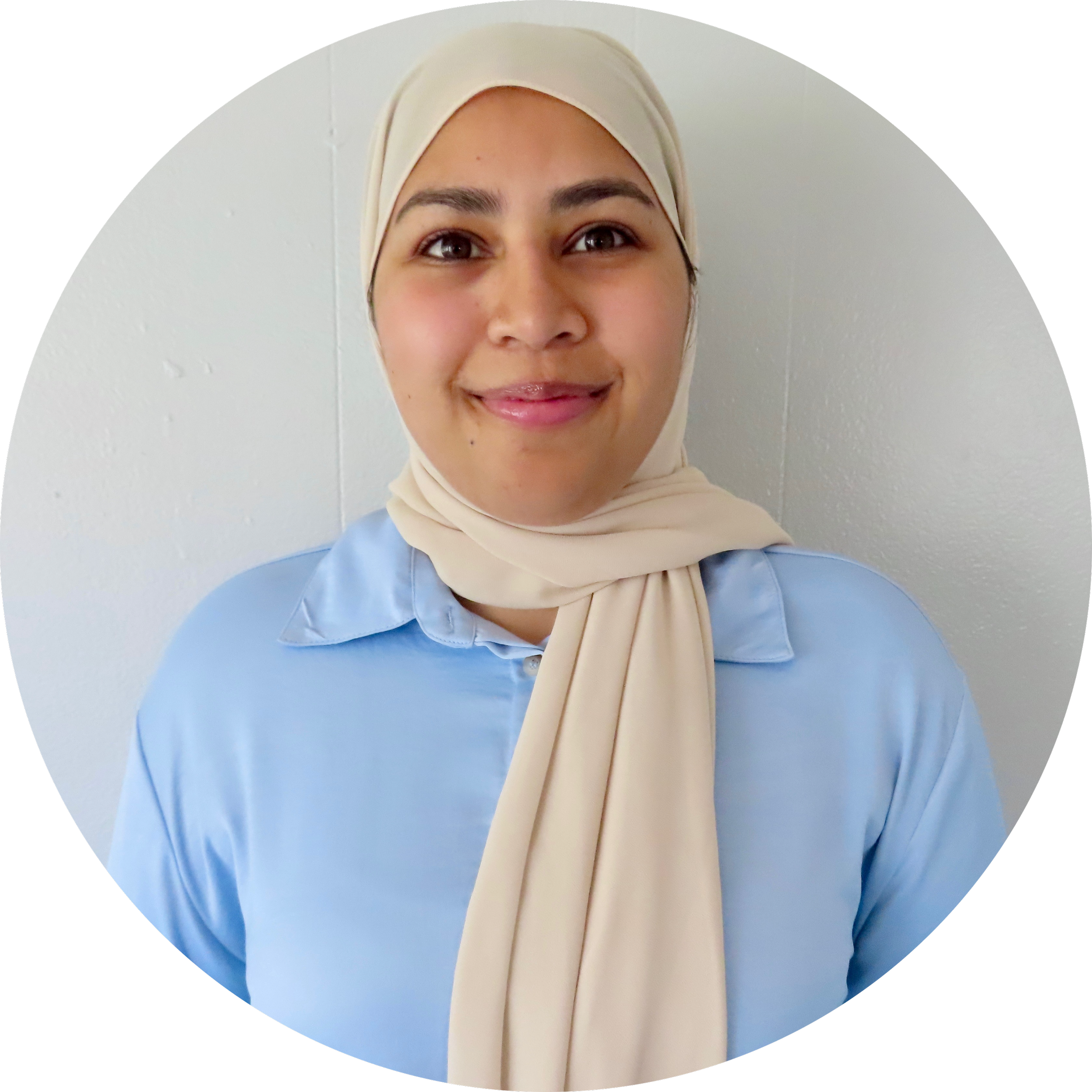 Close-up of a young woman wearing a beige hijab and blue shirt, smiling against a light-colored wall background.