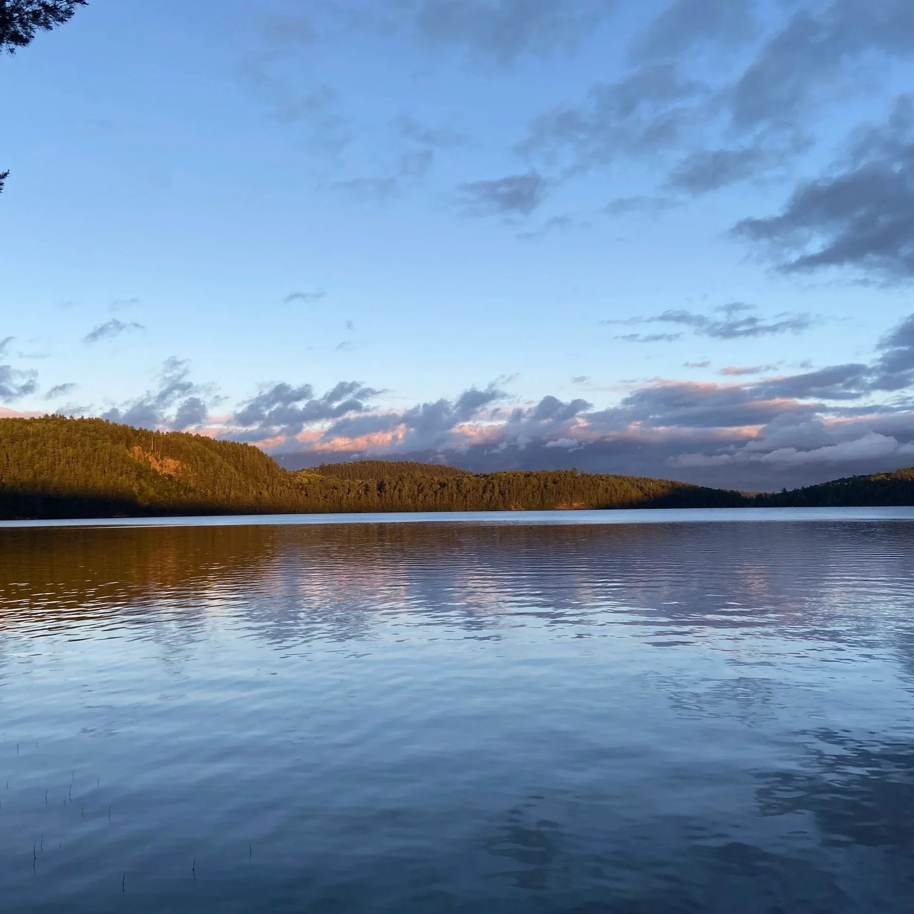 A calm body of water reflecting a sky with scattered clouds, with a tree-covered hill on the left and below the clouds at sunset or sunrise.
