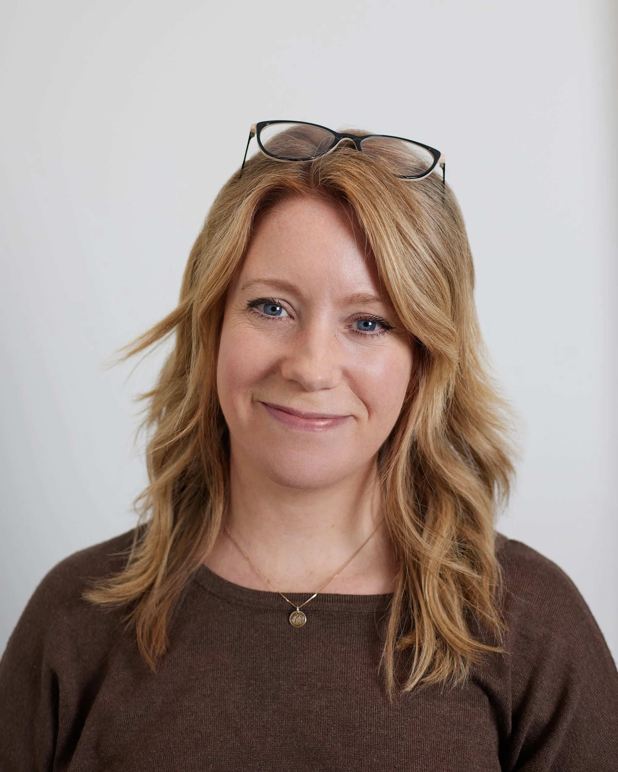Close-up of a smiling woman with blonde hair, wearing glasses on her head, a brown sweater, and a gold necklace with a round pendant, against a plain light gray background.