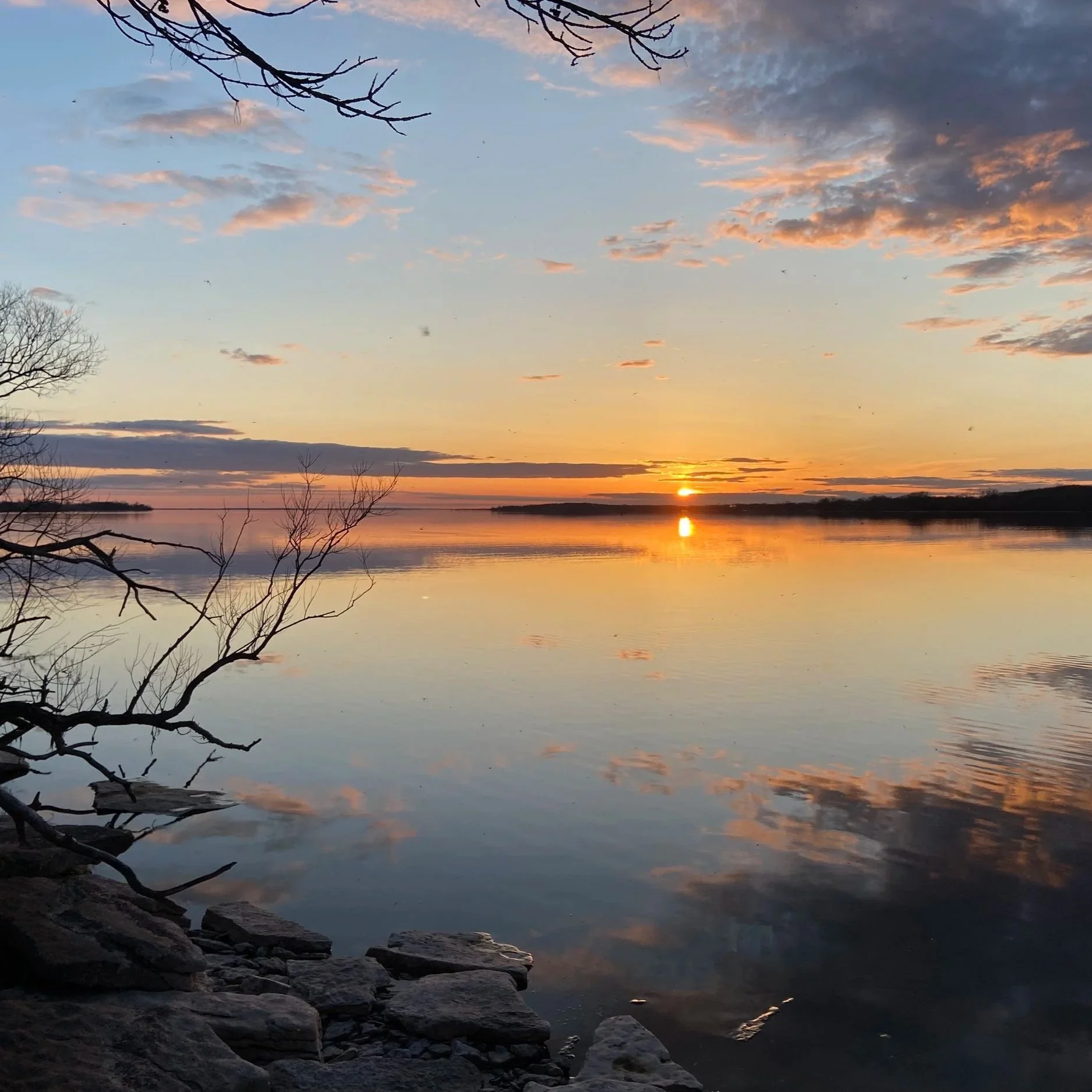 A scenic view of a sunset over a body of water with clouds in the sky. Bare tree branches are in the foreground, and the water reflects the sunset and clouds above.