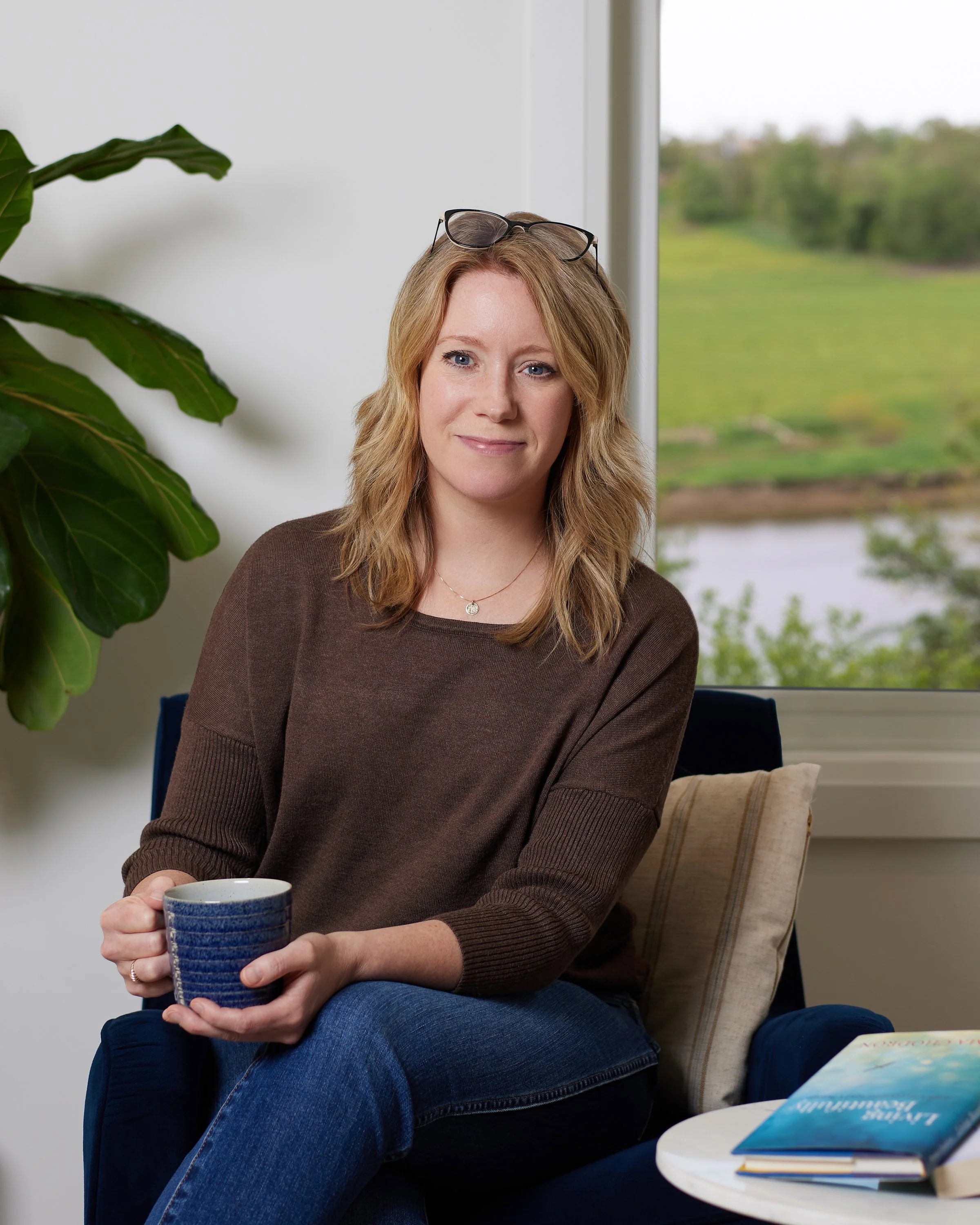 A woman with light brown hair, wearing glasses on her head and a brown sweater, sitting by a window with greenery outside, holding a blue and white striped mug, with books on a small table nearby.