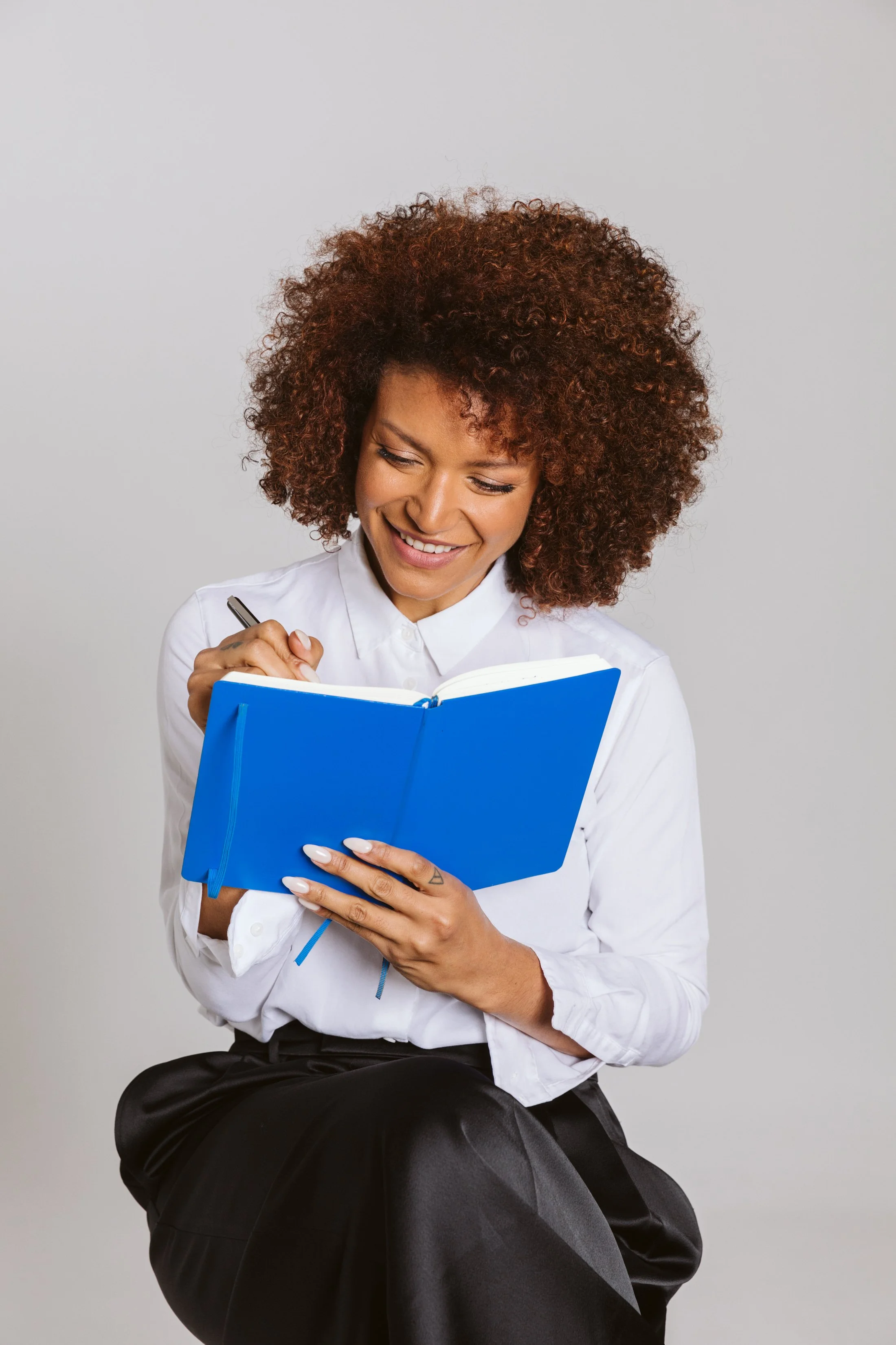 A woman with curly brown hair, wearing a white shirt, sitting on a black chair, holding and writing in a blue notebook, smiling while looking at it against a grey background.