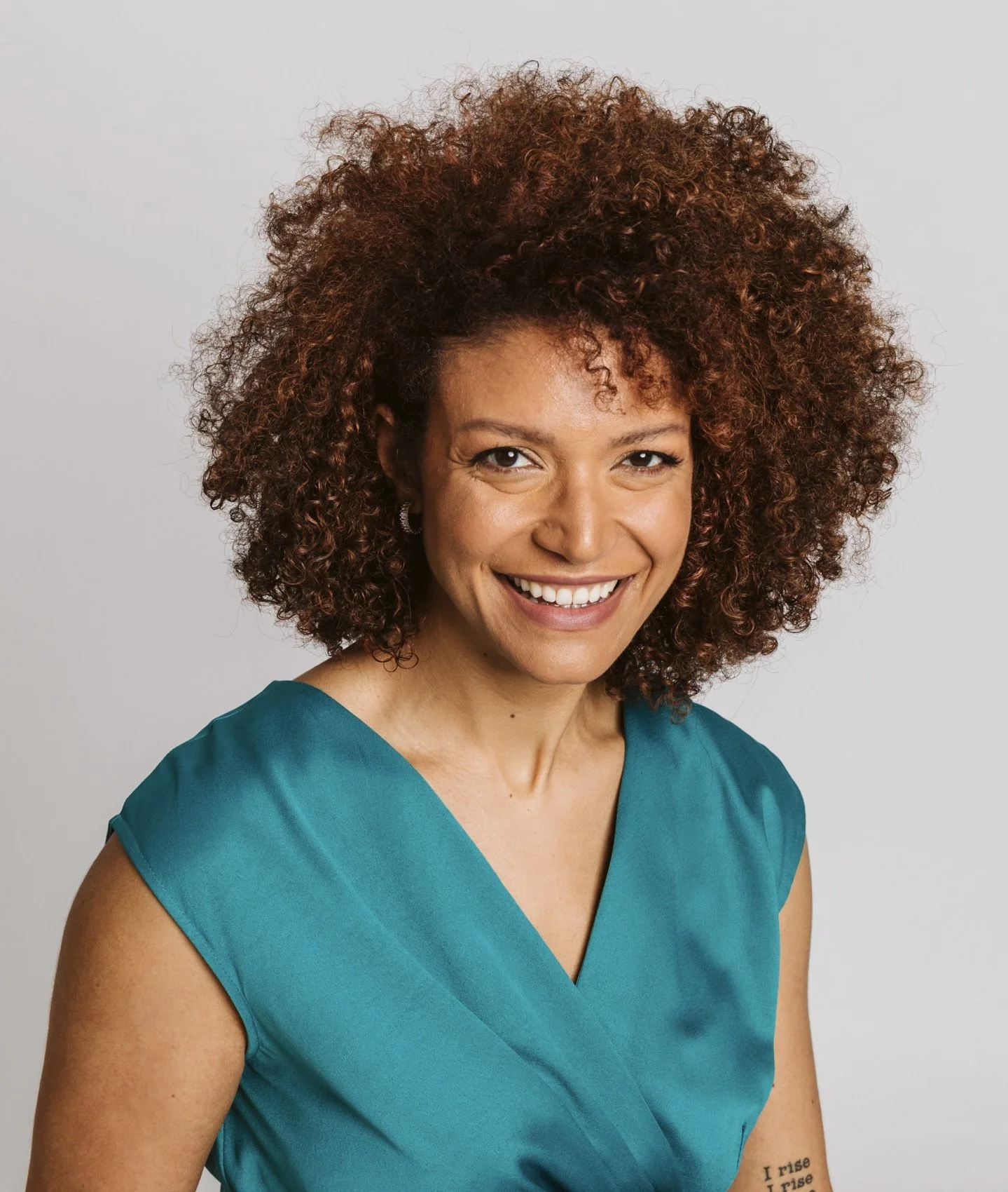 Portrait of a woman with curly brown hair, smiling, wearing a teal dress, against a plain gray background.