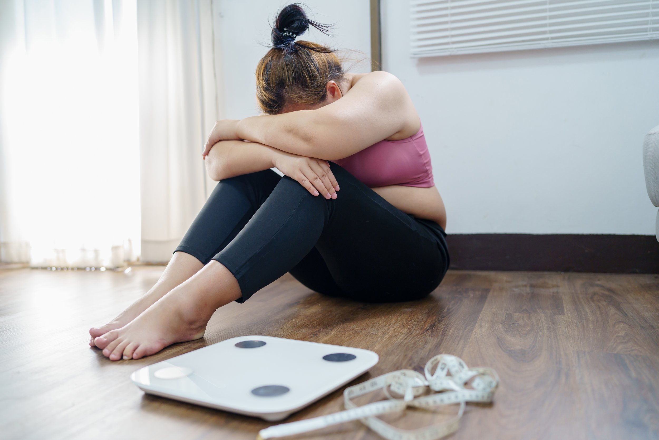 A woman sitting on a wooden floor with her knees drawn to her chest, hugging her legs, and her head resting on her knees. She appears to be upset or distressed. There are measuring tapes and a weighing scale visible on the floor in front of her, with blinds and curtains in the background.