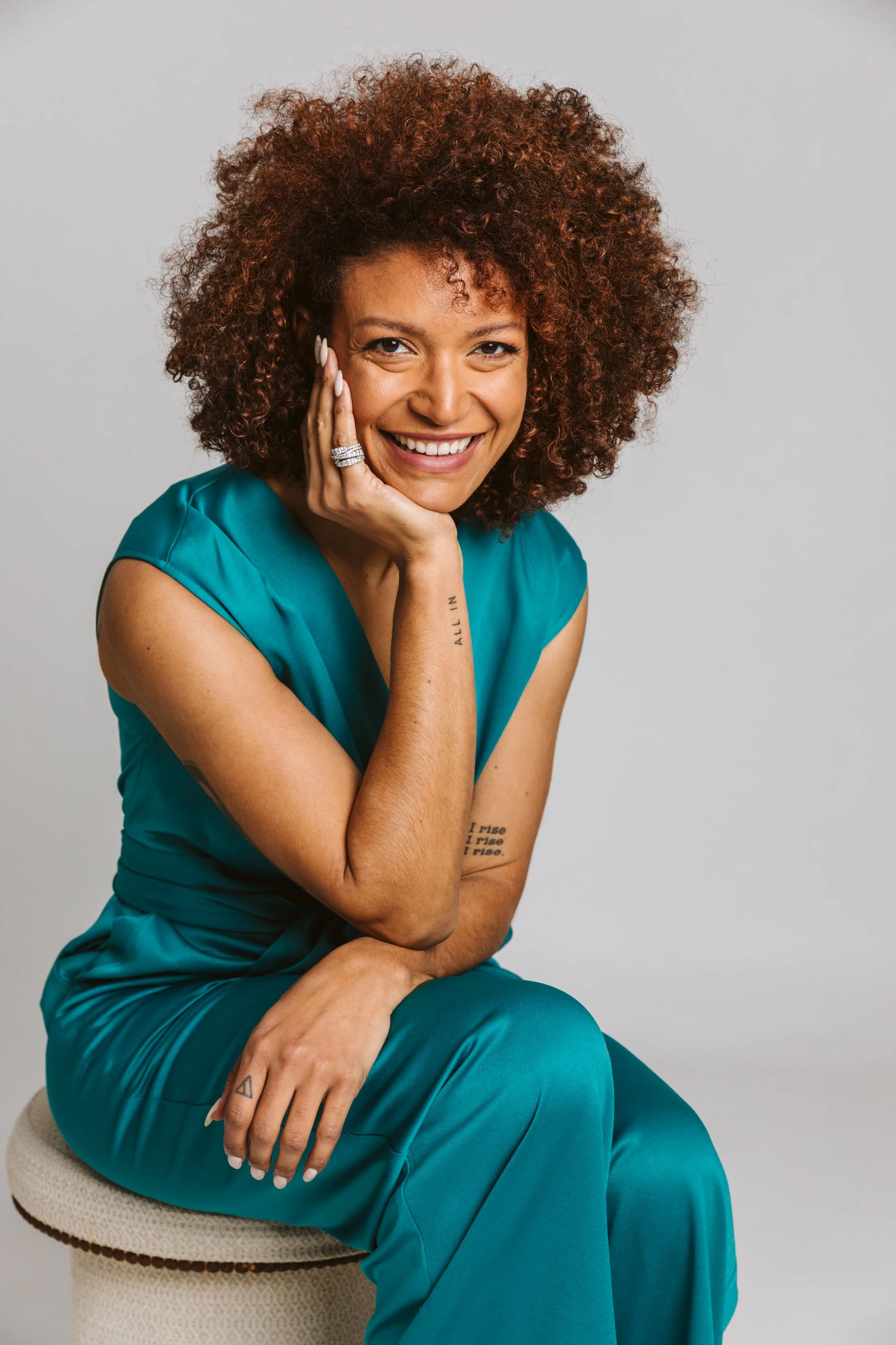 A woman with curly hair wearing blue scrubs sitting on a beige stool, smiling with her hand resting on her face.