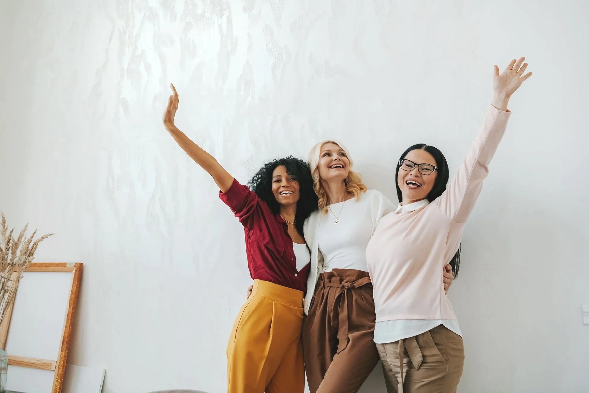 Three women standing together indoors, smiling, with arms raised in celebration. The background is a plain white wall with a framed picture leaning against it.