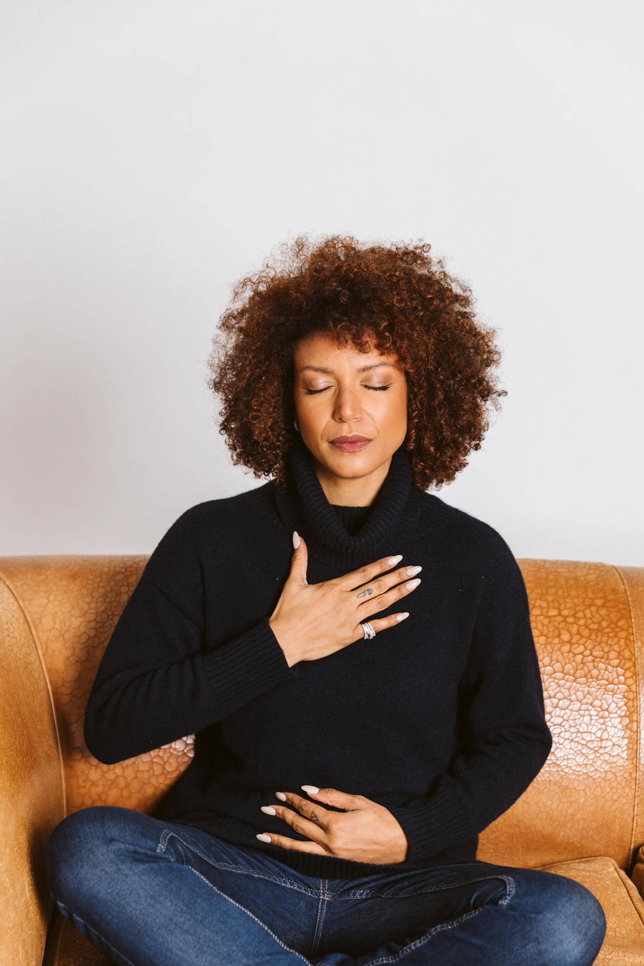 A woman with curly hair and medium skin tone sitting on a beige textured armchair with her eyes closed, wearing a black turtleneck sweater and blue jeans, with her left hand placed on her chest and her right hand on her lap.