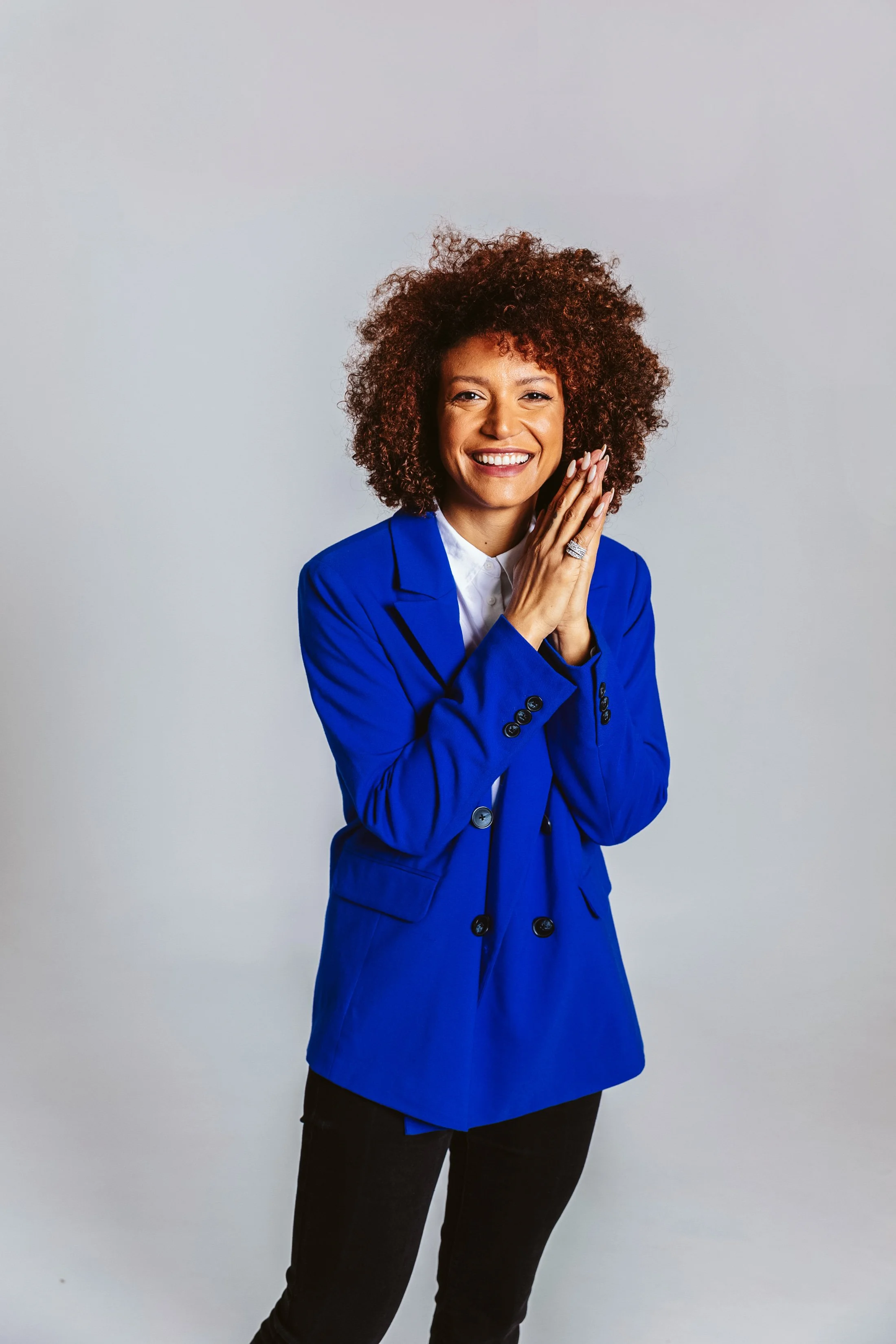 A woman with curly hair wearing a blue blazer and a white blouse, smiling and holding her hands together.