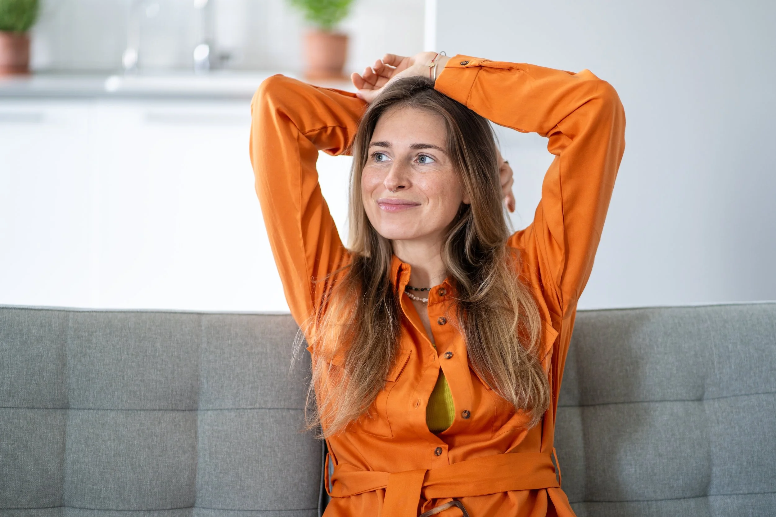 Young woman with long hair and a septum piercing, wearing an orange outfit, sitting on a gray sofa with her arms raised behind her head, smiling and looking to the side, in a bright room with large windows and potted plants in the background.