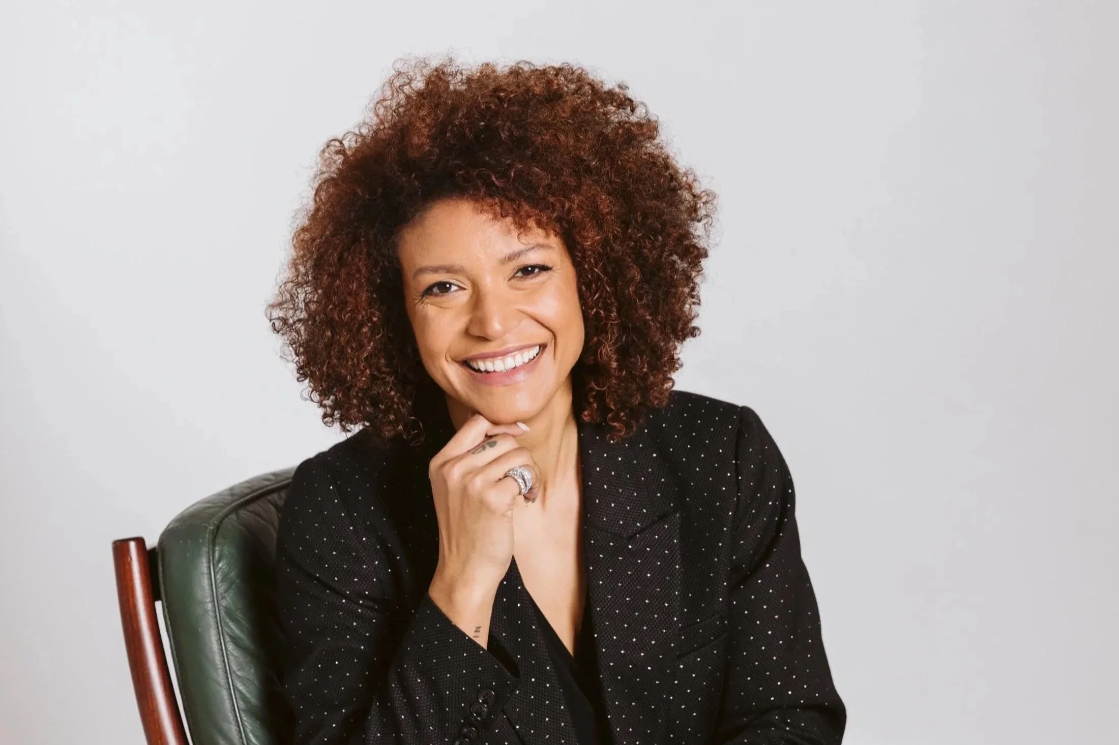 A smiling woman with curly reddish-brown hair, wearing a black polka dot blazer, sitting on a green chair against a plain white background.