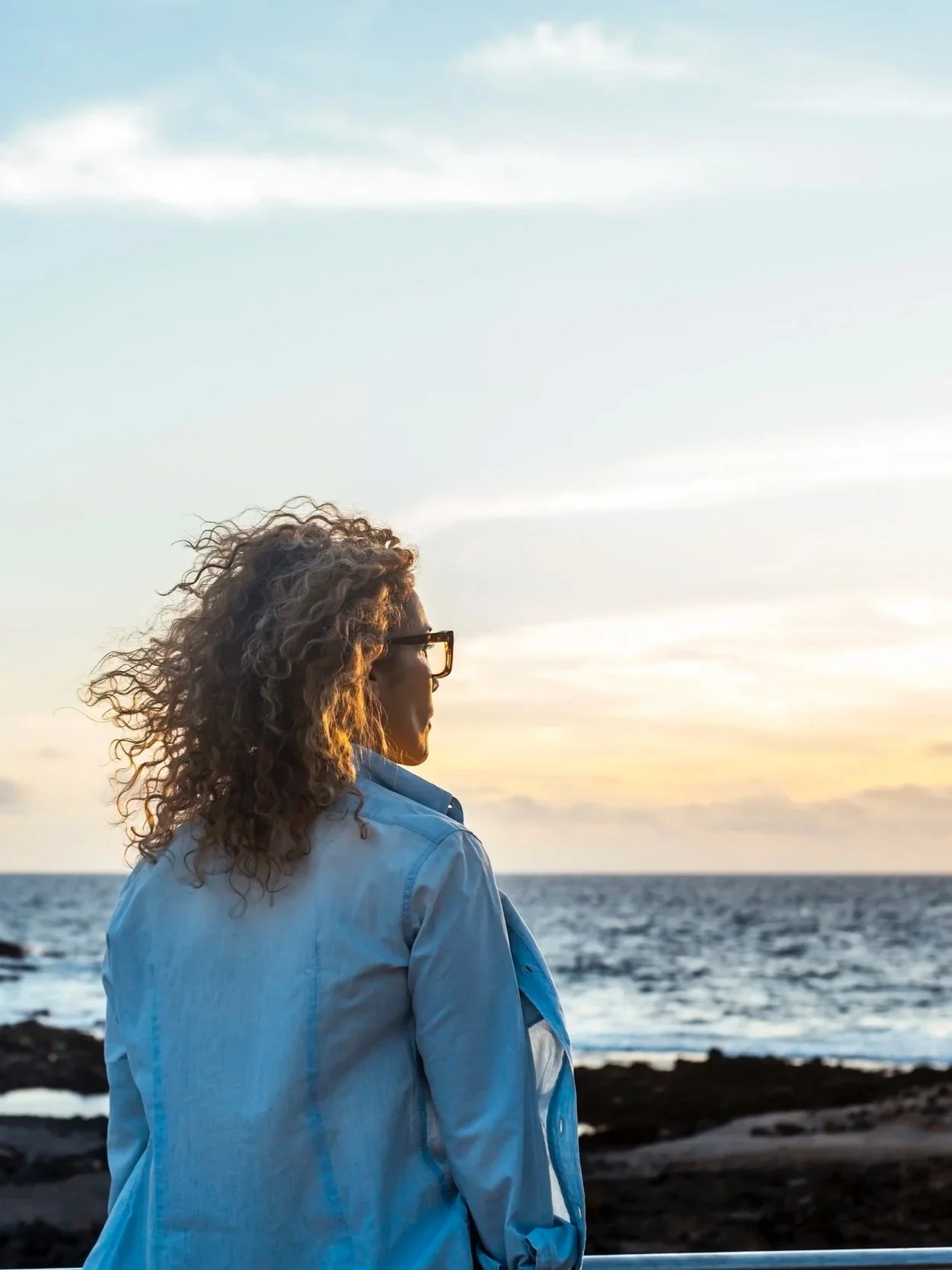 A woman with curly hair and glasses wearing a light blue shirt looking at the ocean during sunset.