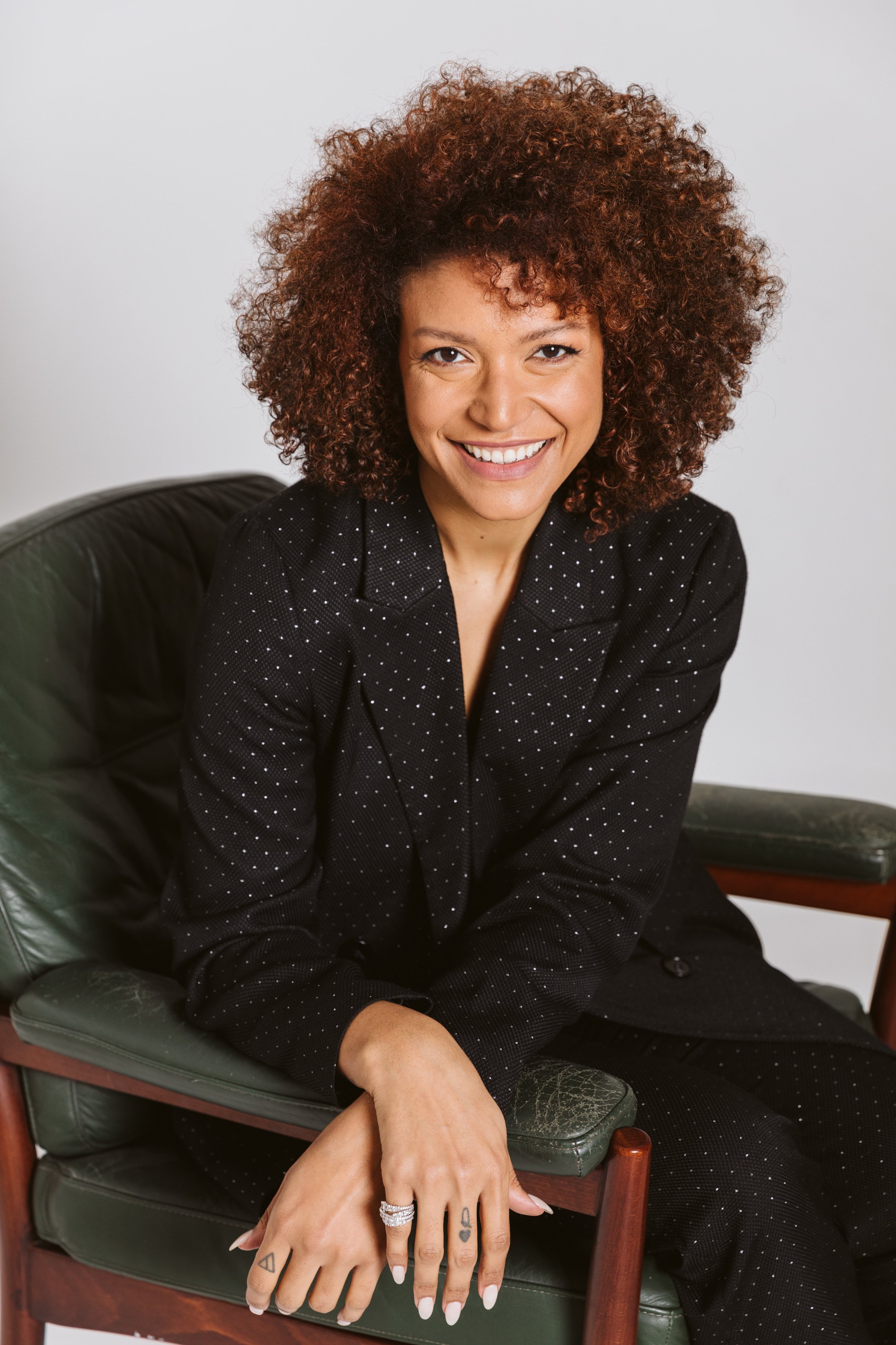 A woman with curly hair sits on a green armchair, smiling at the camera. She wears a black blazer with white polka dots and has rings on her fingers.
