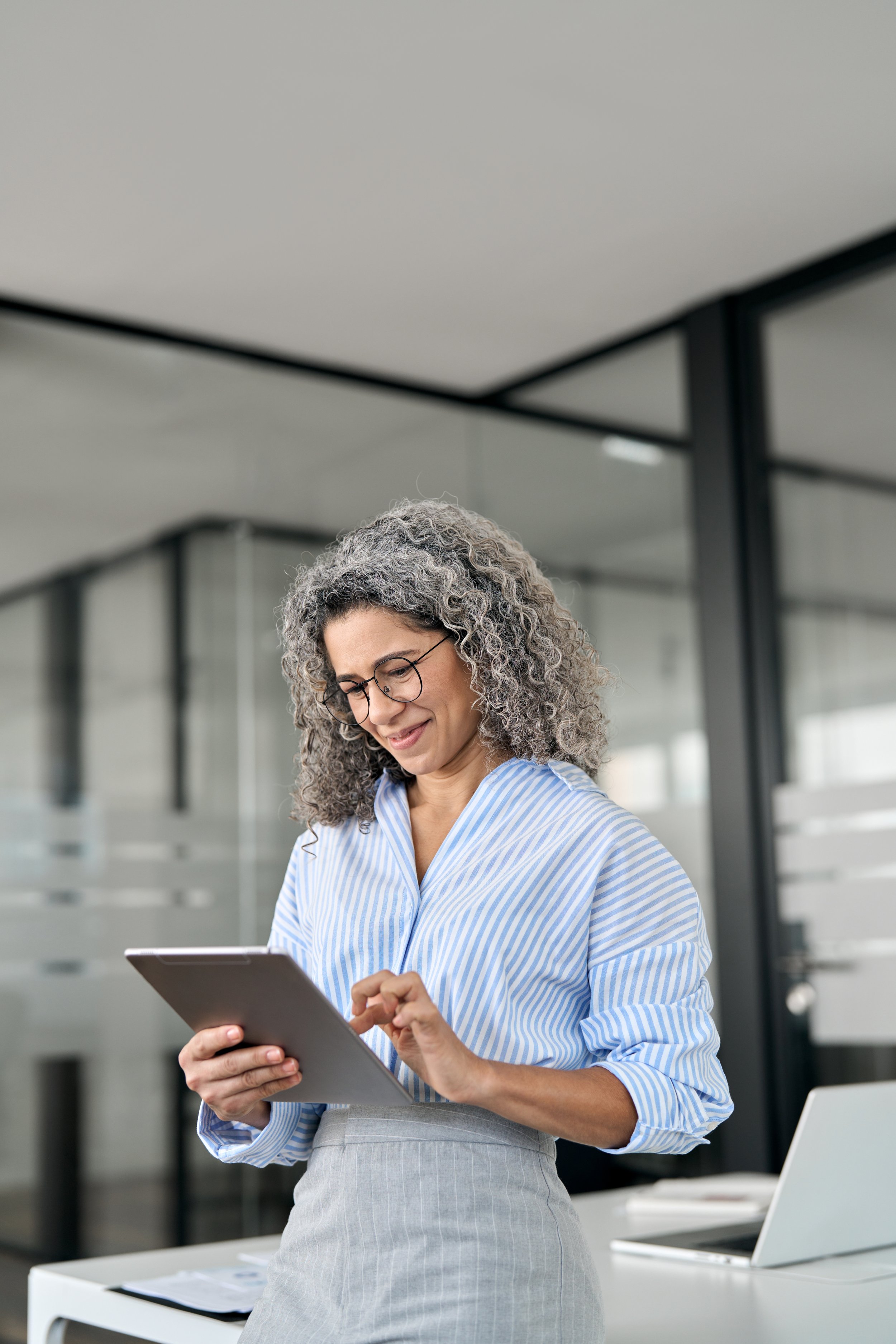 Woman with curly gray hair and glasses wearing a striped blue and white blouse, standing in a modern office, looking at a tablet.