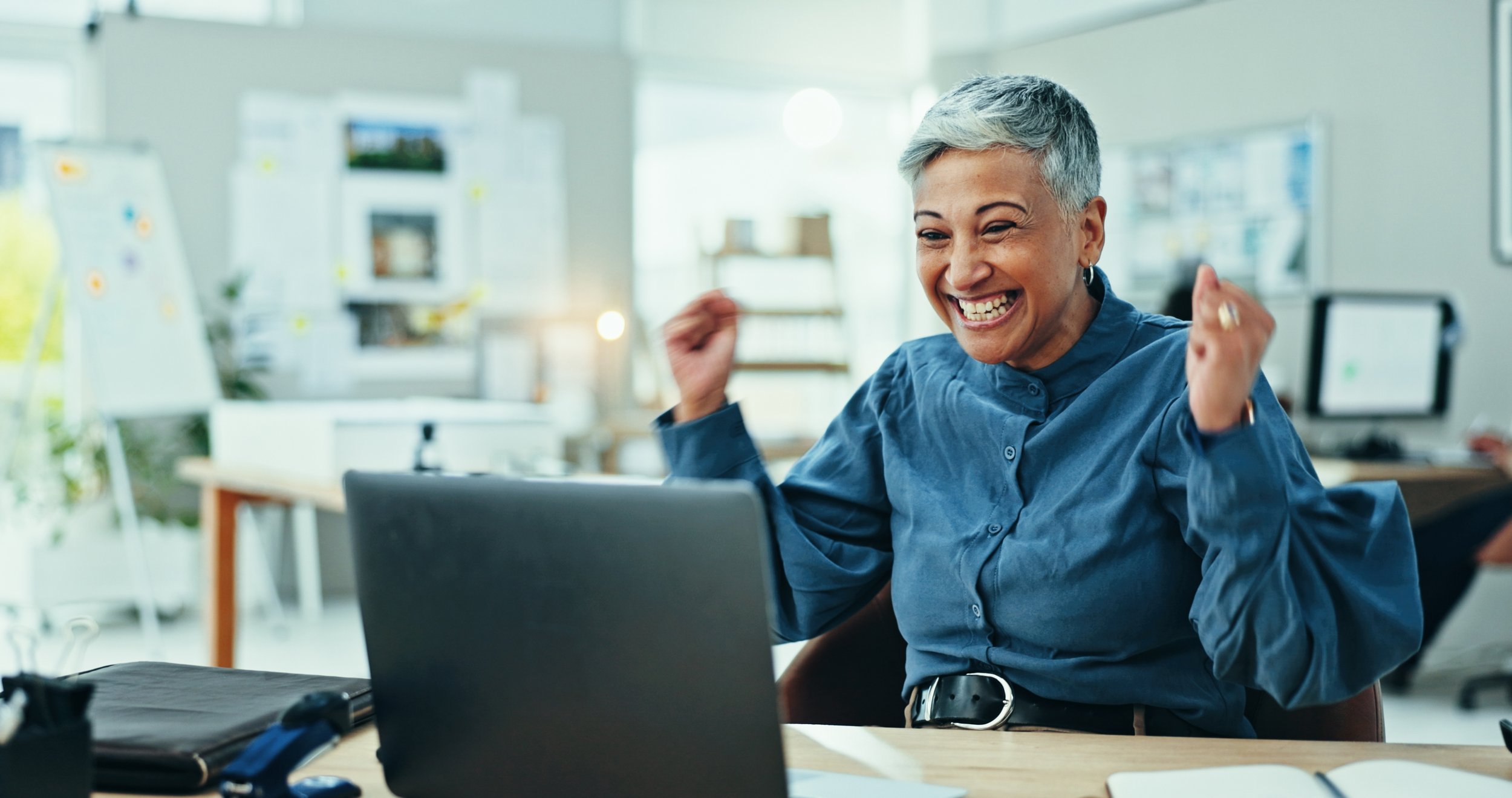 A woman with short gray hair wearing a blue shirt, smiling and celebrating while sitting at a desk in an office environment with a laptop and various office supplies.