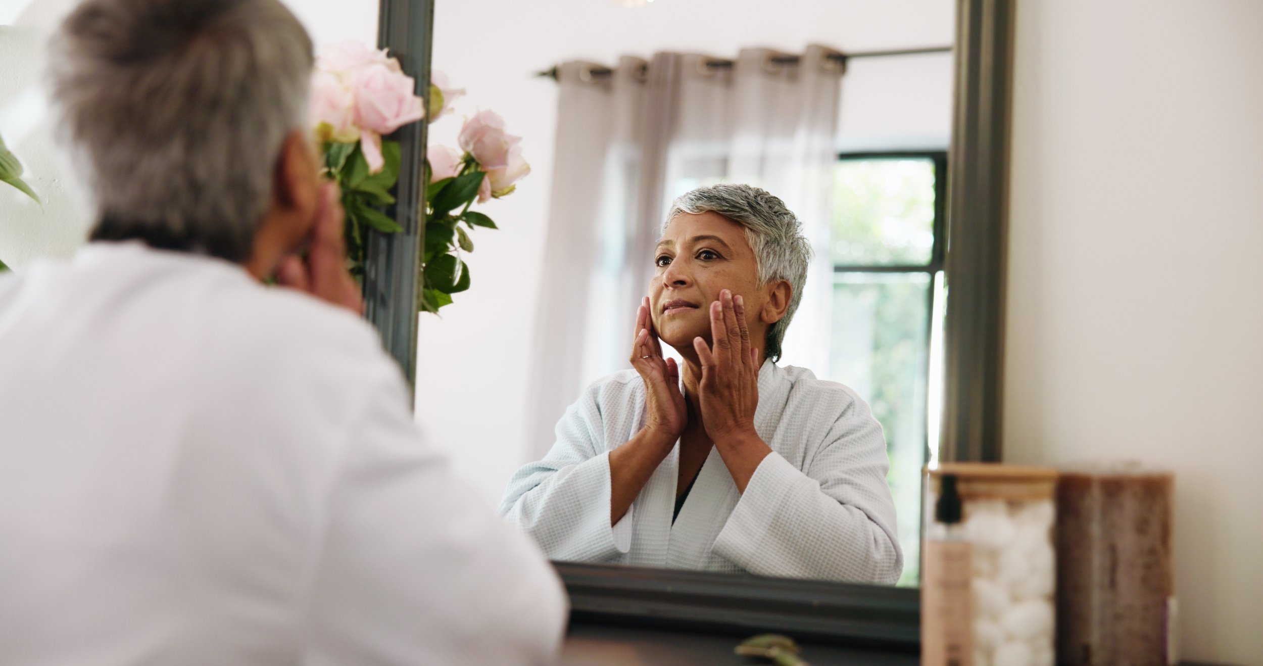 An elderly woman in a white bathrobe looks at herself in a mirror while touching her face, with a man observing her reflection, both in a room with natural light and pink flowers on the window sill.