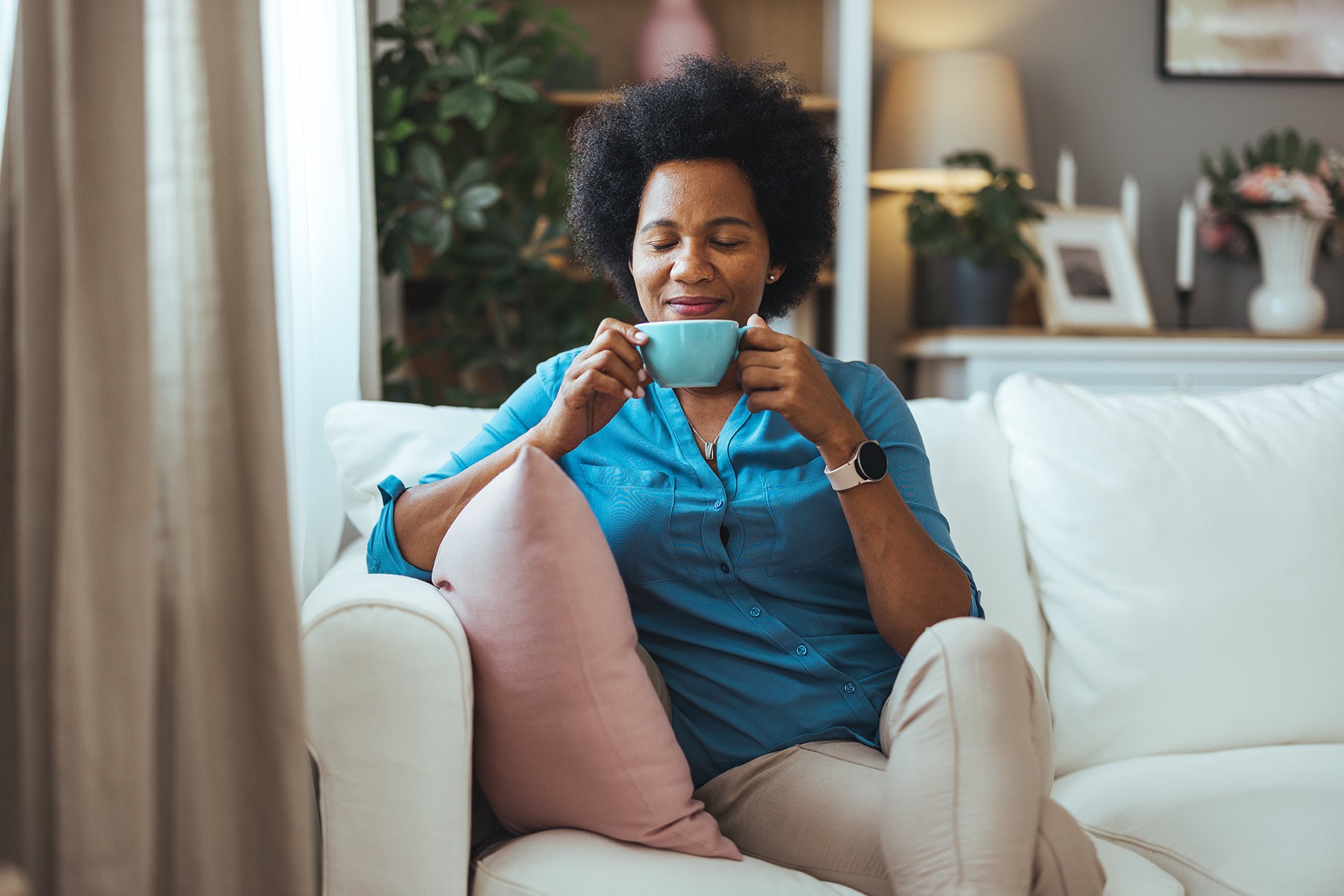 A woman with curly black hair, wearing a blue blouse, sitting on a cream-colored sofa, holding a blue mug, smiling with eyes closed in a cozy living room with plants and framed photos in the background.