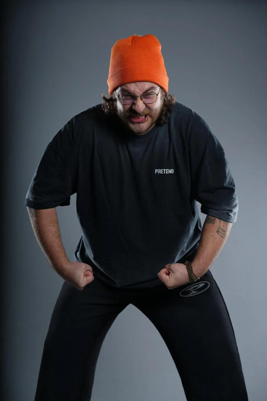 Man wearing an orange beanie and glasses flexing his arms, with a serious expression, against a gray background.