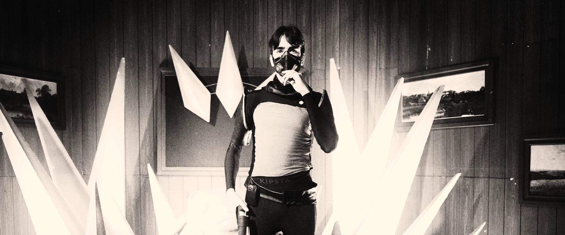 Person wearing a mask and athletic clothing, standing indoors with pointed white sculpture pieces around them, and framed landscape paintings on the wooden-paneled walls.