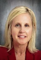 Portrait of a woman with shoulder-length blonde hair, wearing a red blazer, against a dark, blurred background.