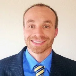 A man with short light brown hair and a beard, wearing a dark pinstripe suit, light blue shirt, and a striped tie, smiling against a plain light background.