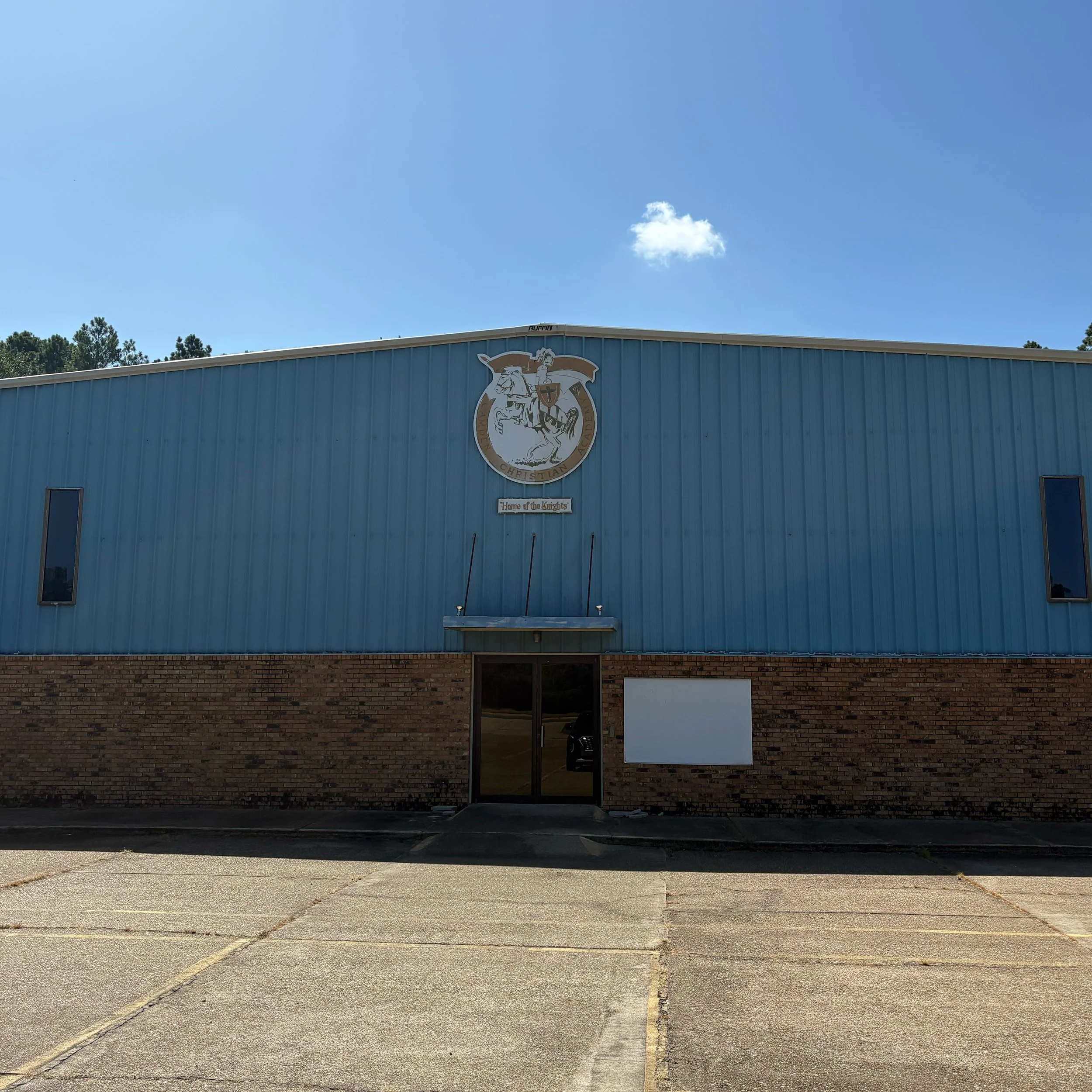 The exterior of a blue building with a logo and a sign that reads 'Home of the Knights'. There is a parking lot in front with marked parking spaces and some small trees. The sky is mostly clear with one small cloud.