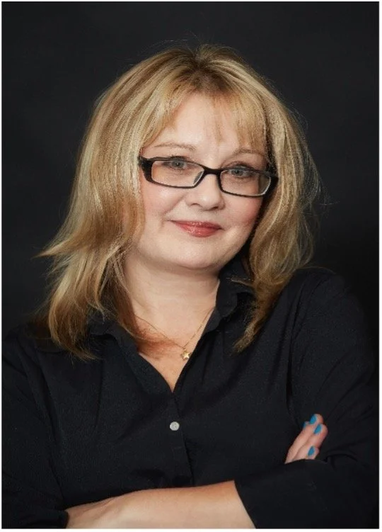 A woman with blonde hair, glasses, and a black shirt posing with her arms crossed against a dark background.