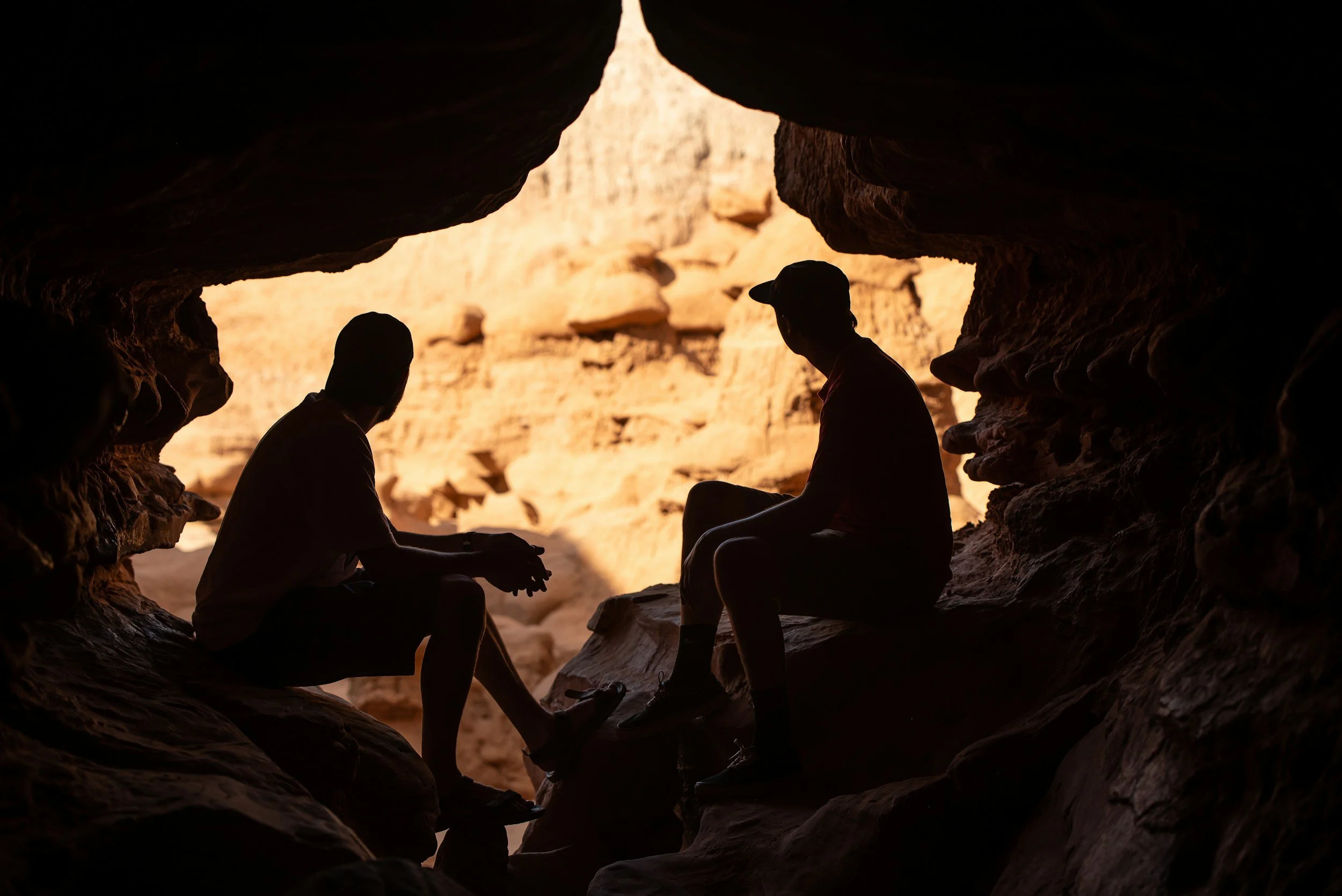 Silhouettes of three people sitting in a rock formation at sunset or sunrise, with a view of a canyon or desert landscape in the background.