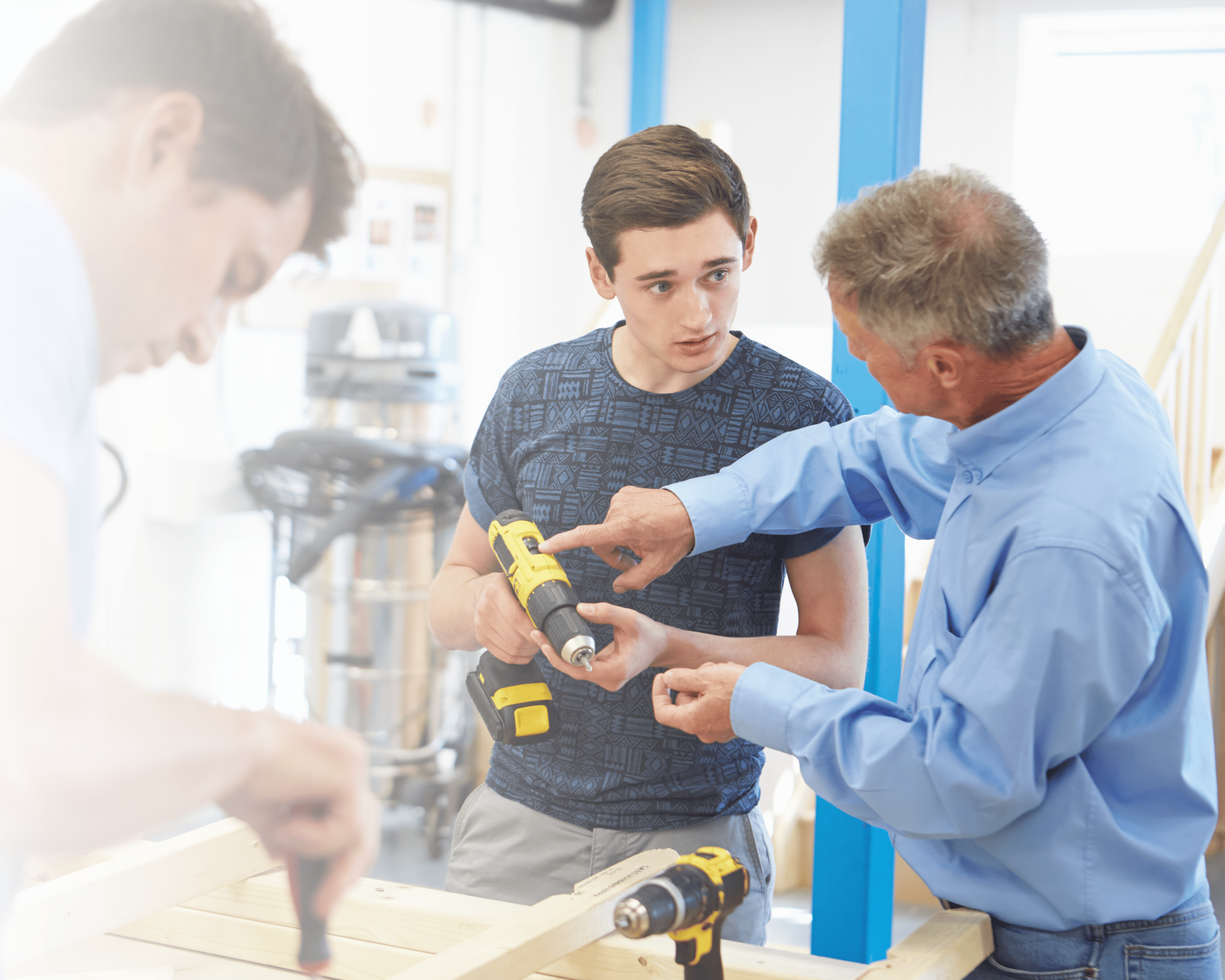 A young man in a patterned blue shirt demonstrates a cordless drill to an older man in a light blue shirt in a bright workshop with blue accents.