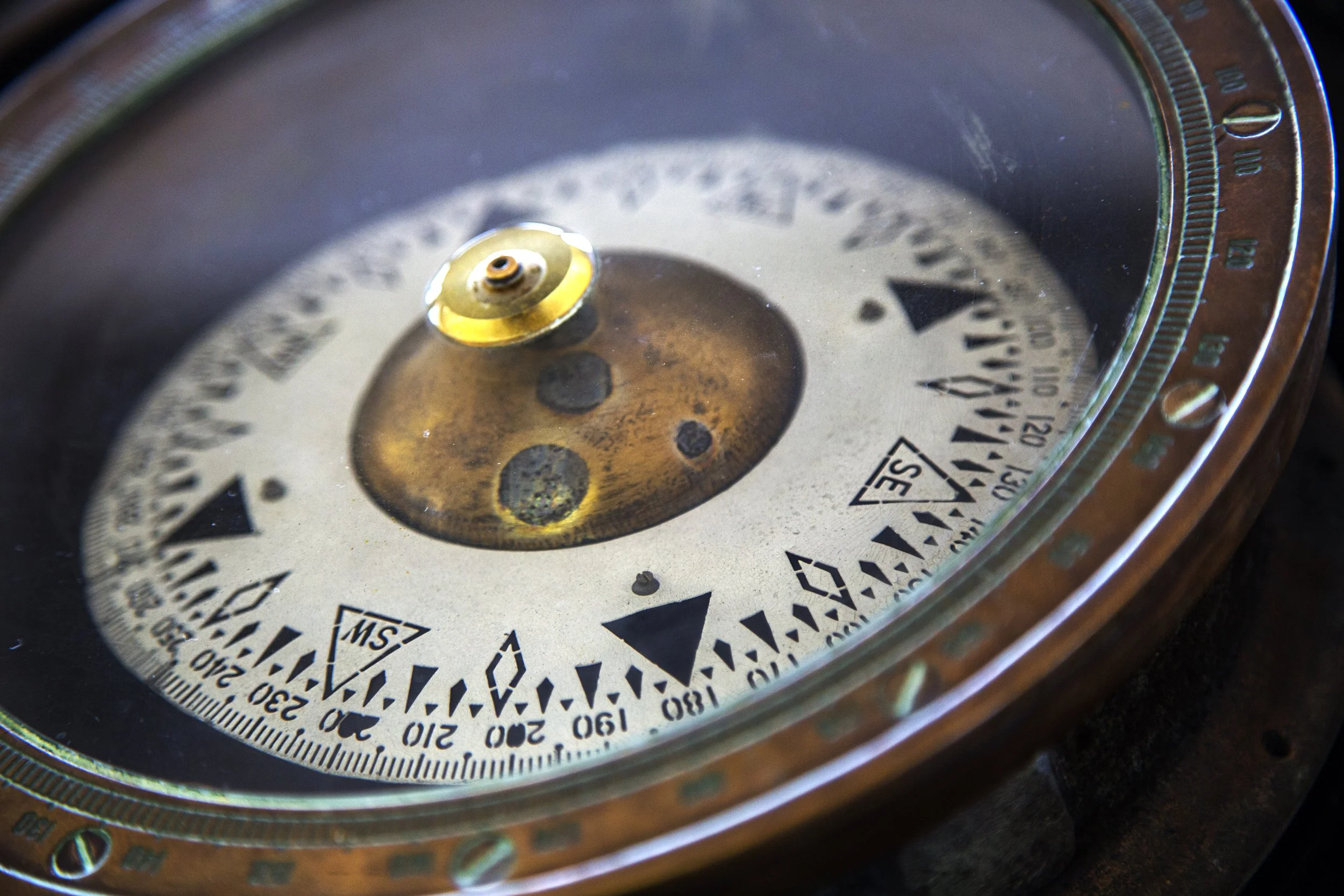 Close-up photo of a compass with a metallic face, showing directional markings and a gold-colored central screw, reflecting light.