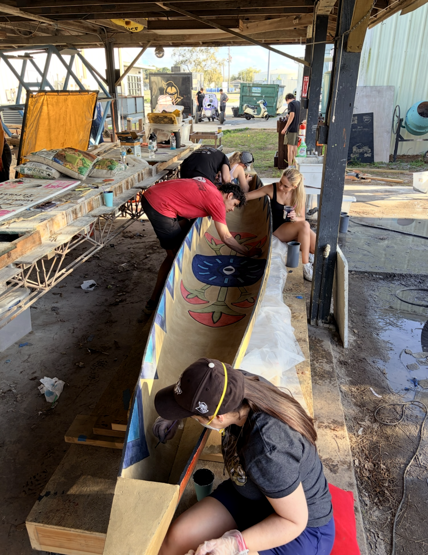 People working on a long painted wooden project in a covered outdoor space, with tools, snacks, and water bottles on the tables, and various objects and scooters outside.