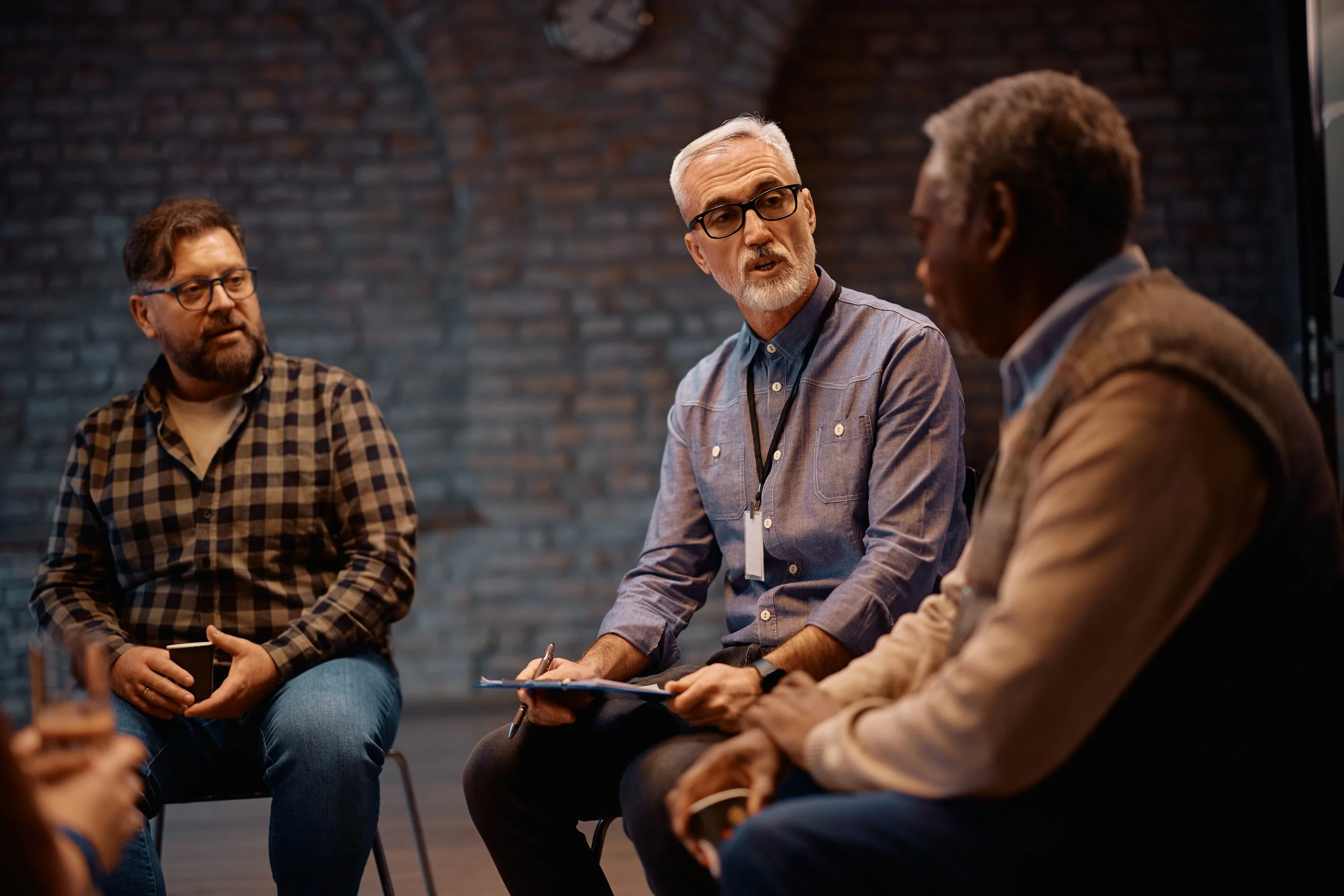 A group of diverse adults engaged in a discussion, sitting in a circle in a room with brick walls.