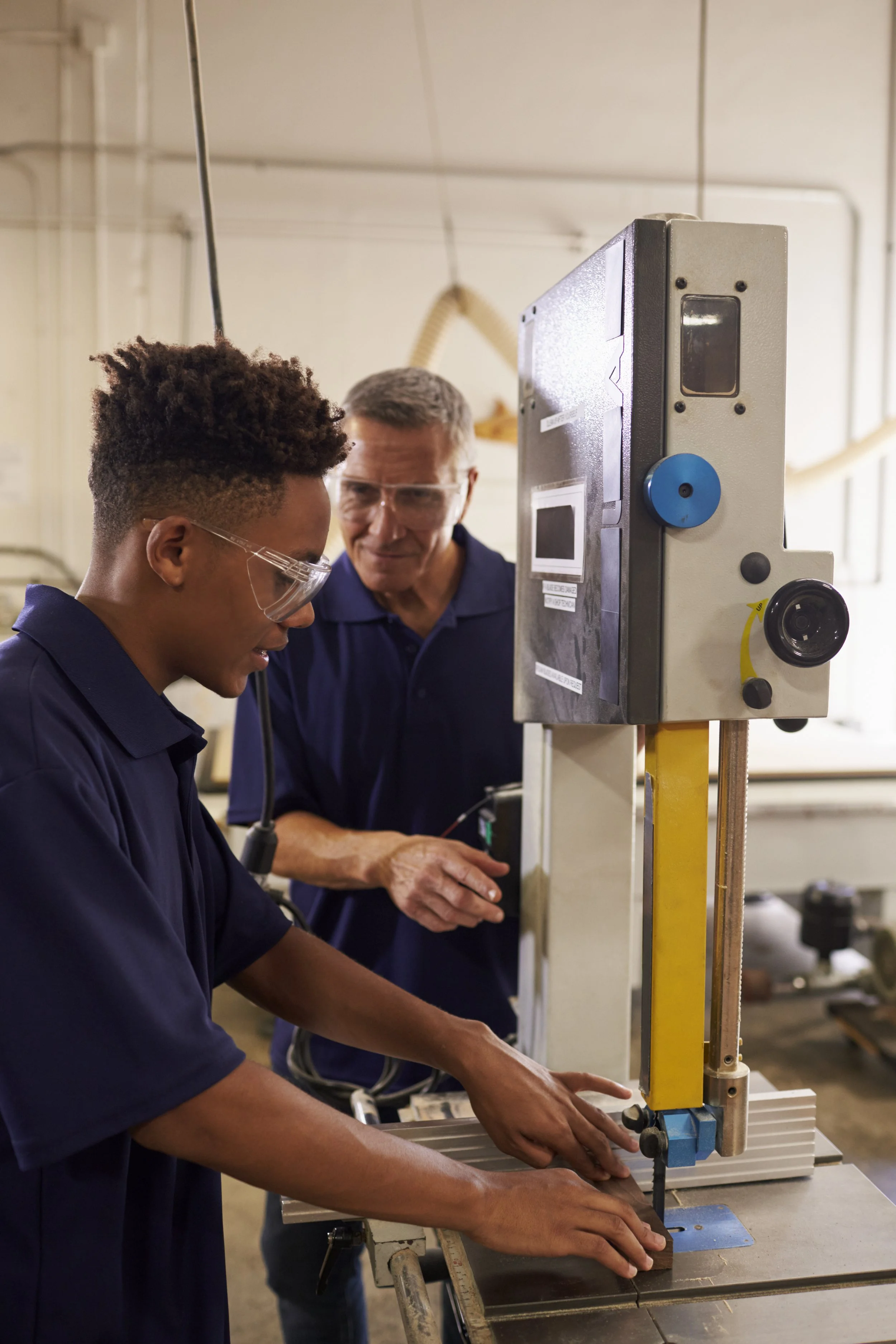 Two men working together on a woodworking or manufacturing project in a workshop. One is wearing safety glasses and the other is looking on. They are handling a machine or tool together.