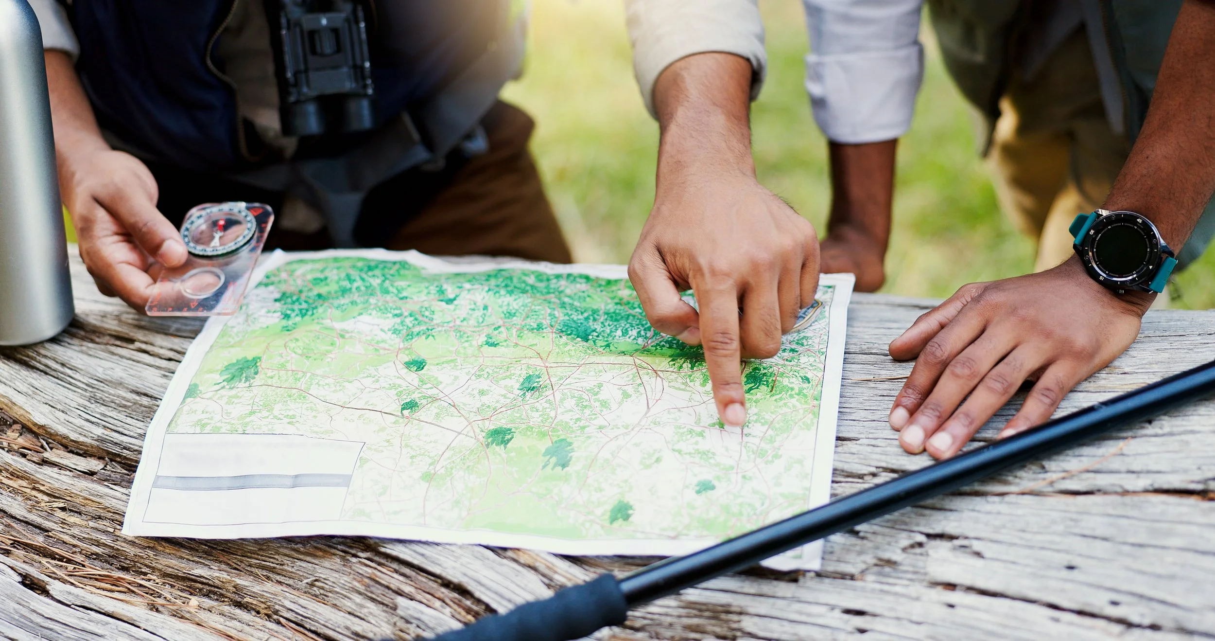 People examining a topographical map outdoors, with one person pointing at it, on a wooden surface with various navigation tools nearby.