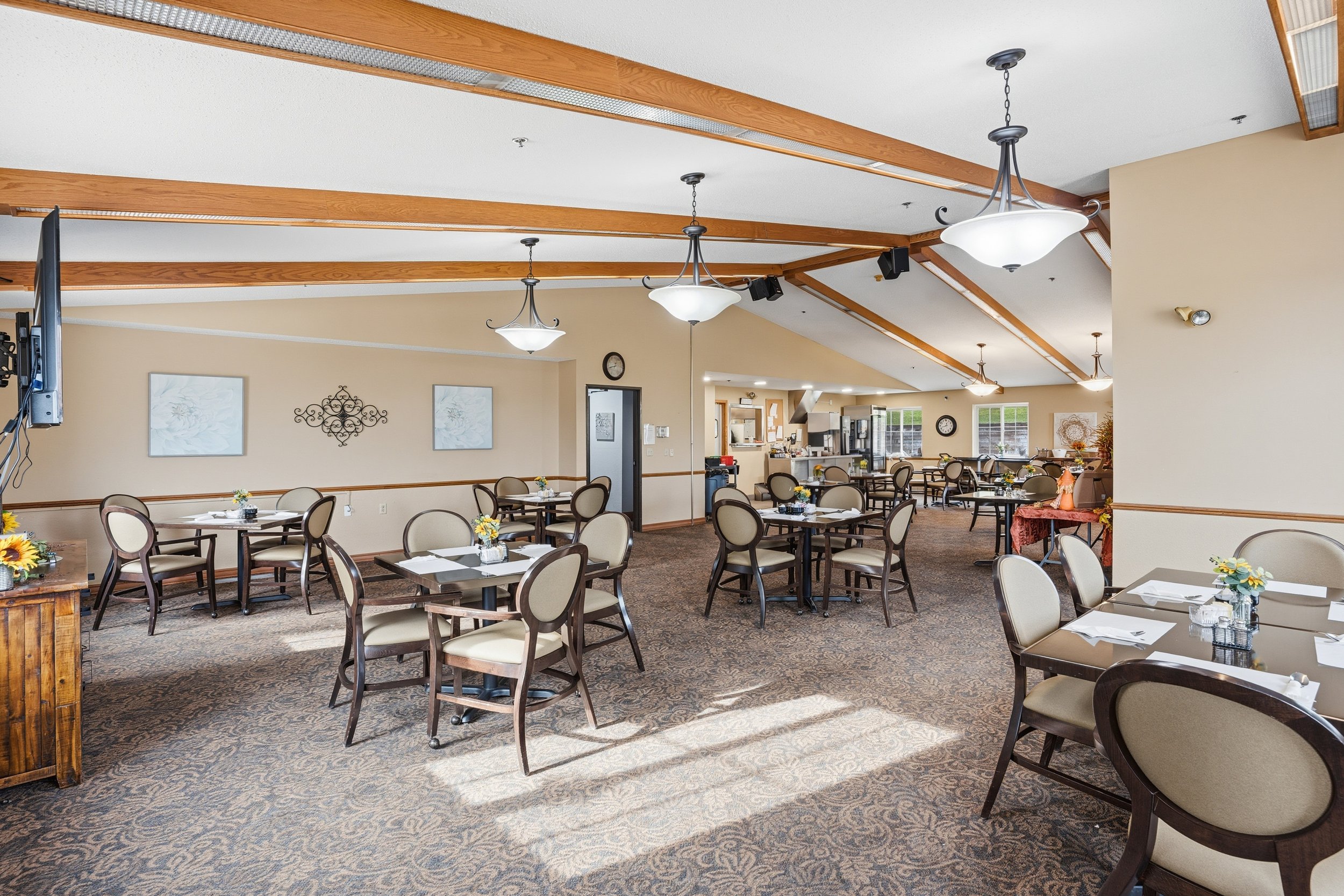A spacious dining area with round tables covered with white napkins and small flower arrangements, surrounded by beige upholstered chairs with dark wood frames, and decorated with sunflowers, wall art, and ceiling lights.