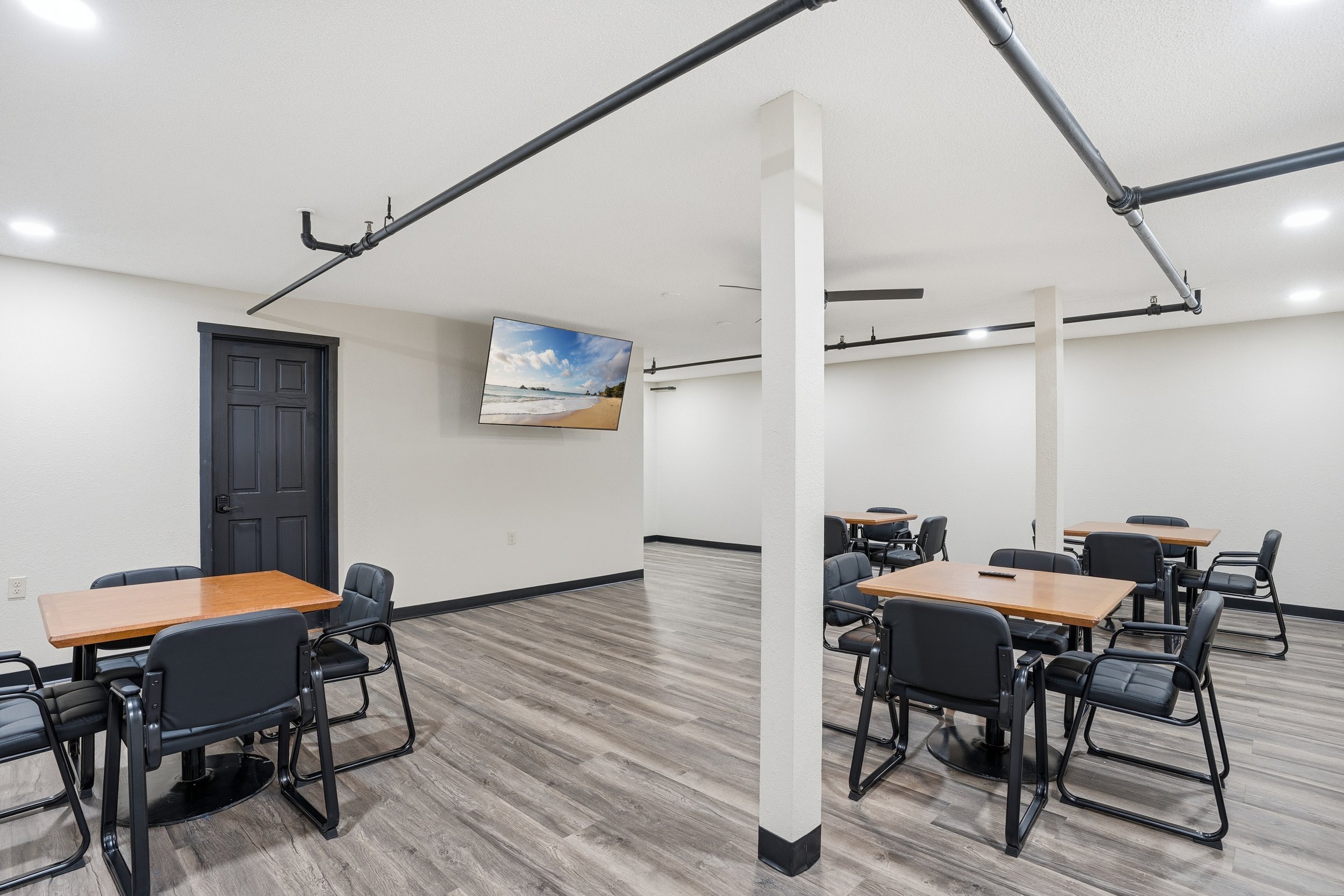 Empty conference or meeting room with multiple tables, black chairs, white walls, a TV screen displaying a beach scene, and black ceiling pipes.