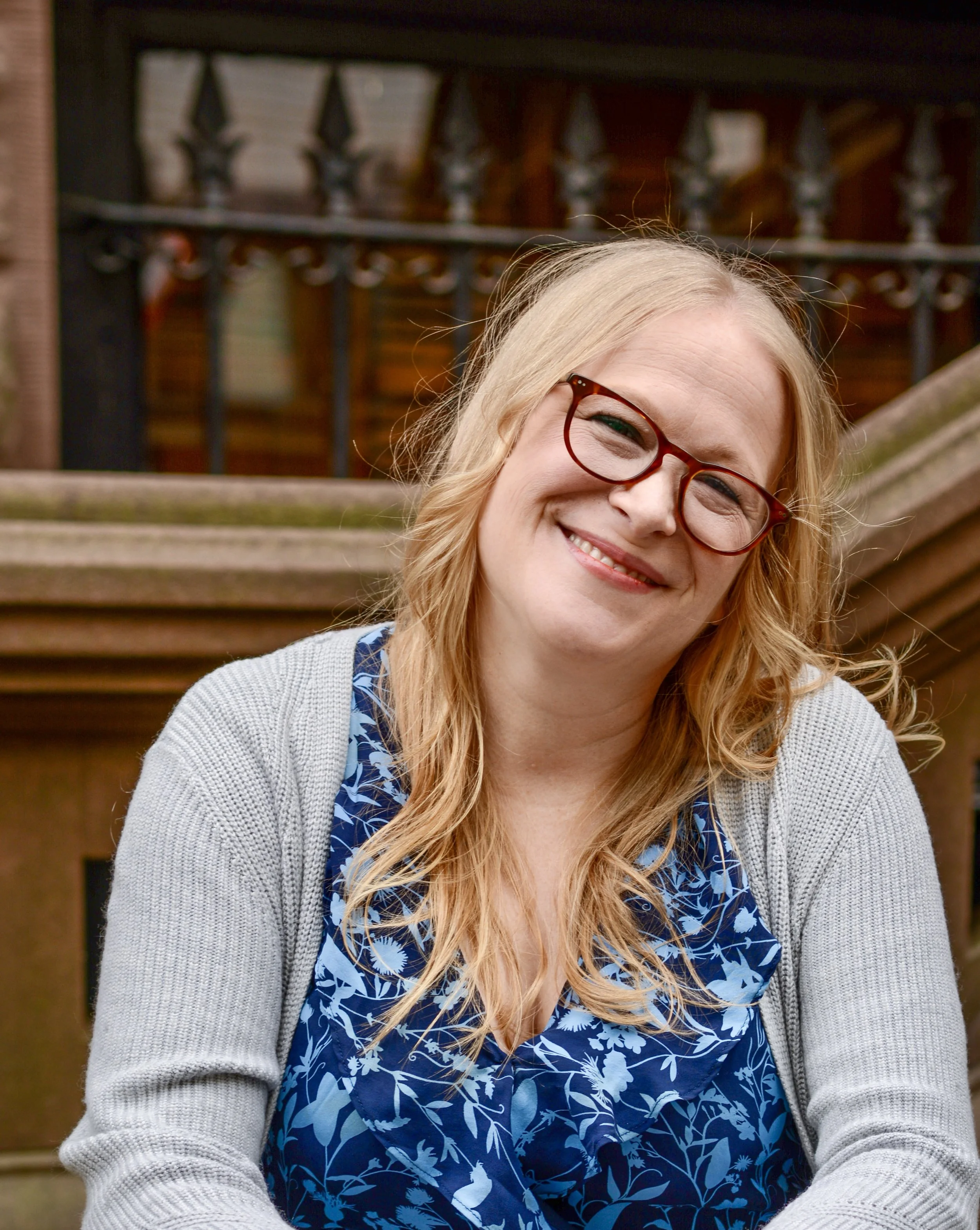 Smiling woman with blond hair and glasses outdoors, sitting on a wooden bench.