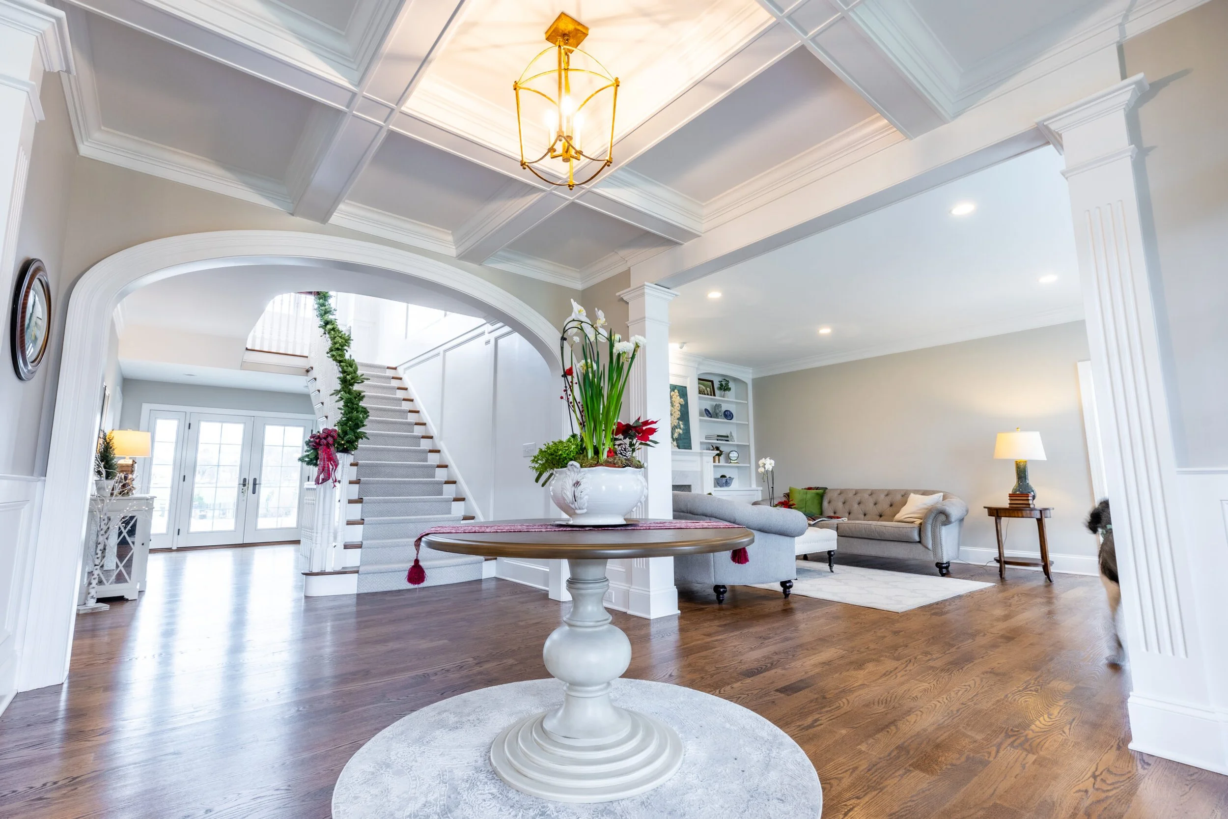 Living room with a white round table, floral centerpiece, taupe sofa, bookshelf, and staircase in the background.